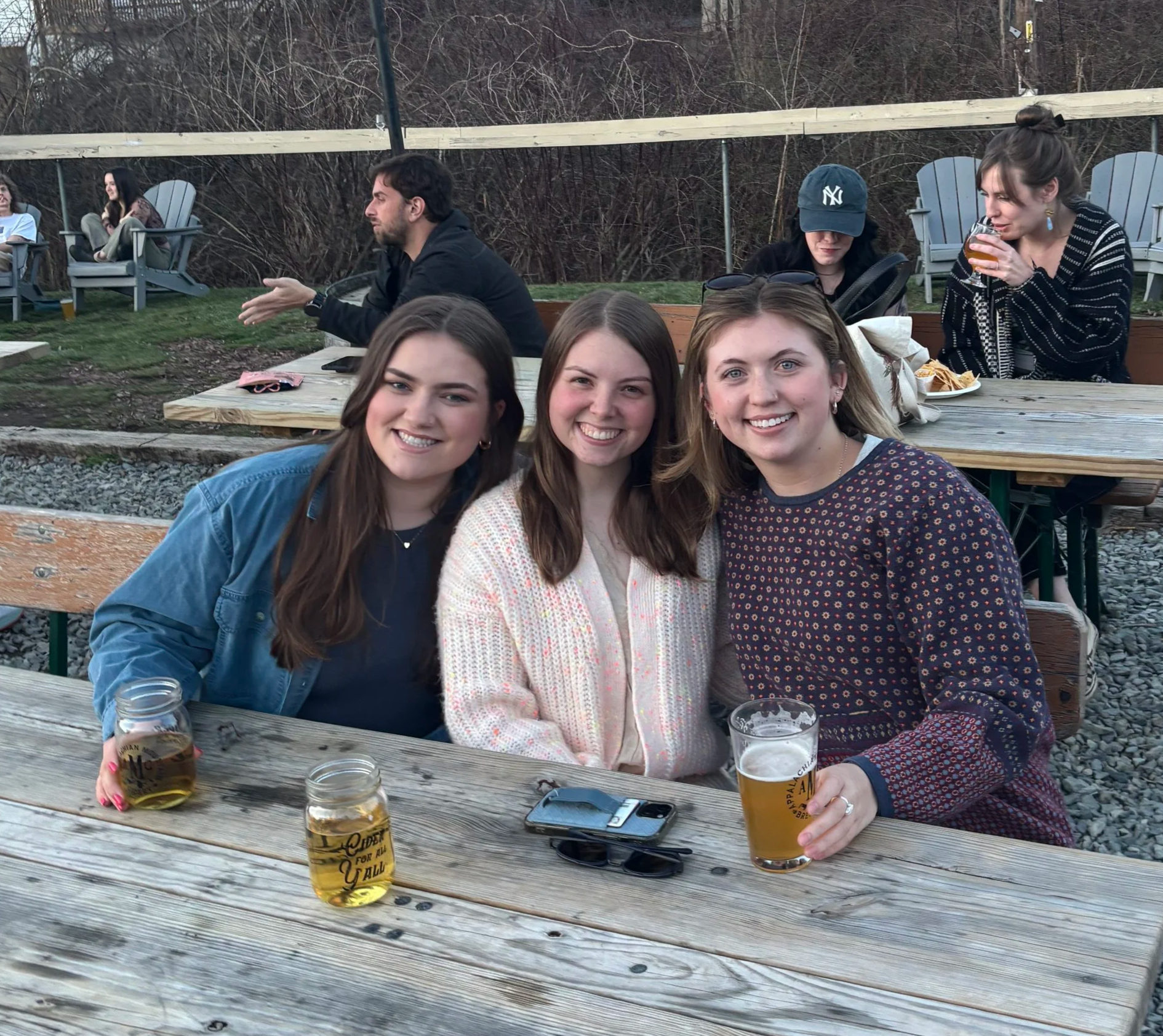 Three young women sitting at a wooden outdoor picnic table, smiling at the camera, with drinks in hand. Behind them, several other people are seated at tables and in chairs, enjoying a social gathering outdoors.