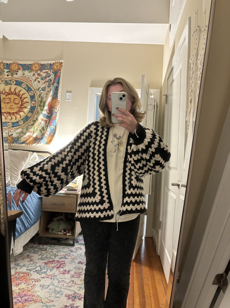 A person with shoulder-length blonde hair taking a mirror selfie in a bedroom, wearing a black and white zigzag patterned jacket and black pants.