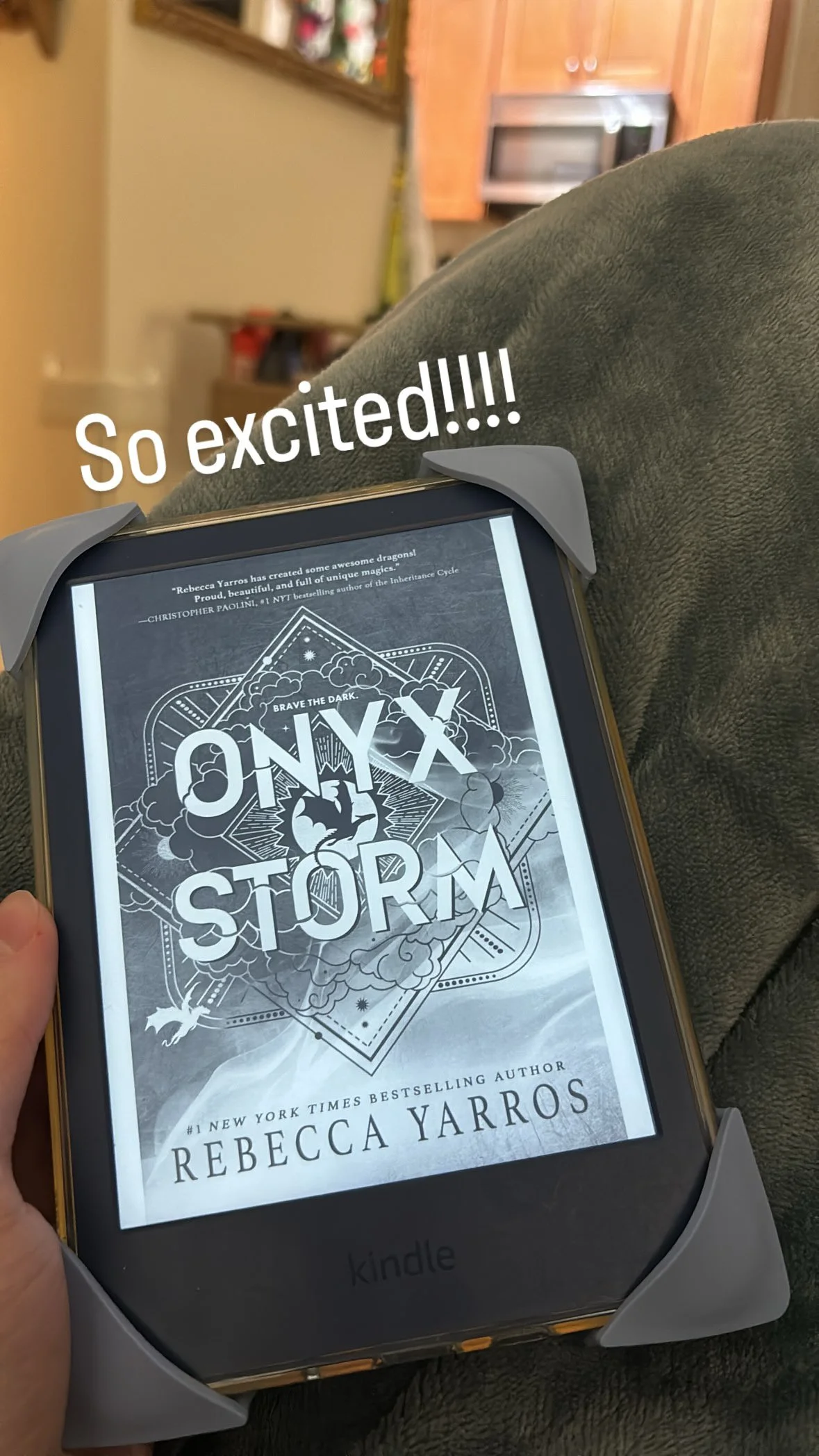 Person sitting on a couch holding a Kindle e-reader with the cover of the book 'Onyx Storm' by Rebecca Yarros visible. The book cover features a dark, stormy sky with clouds, lightning, and a black cat. Text overlay says 'So excited!!!!'.