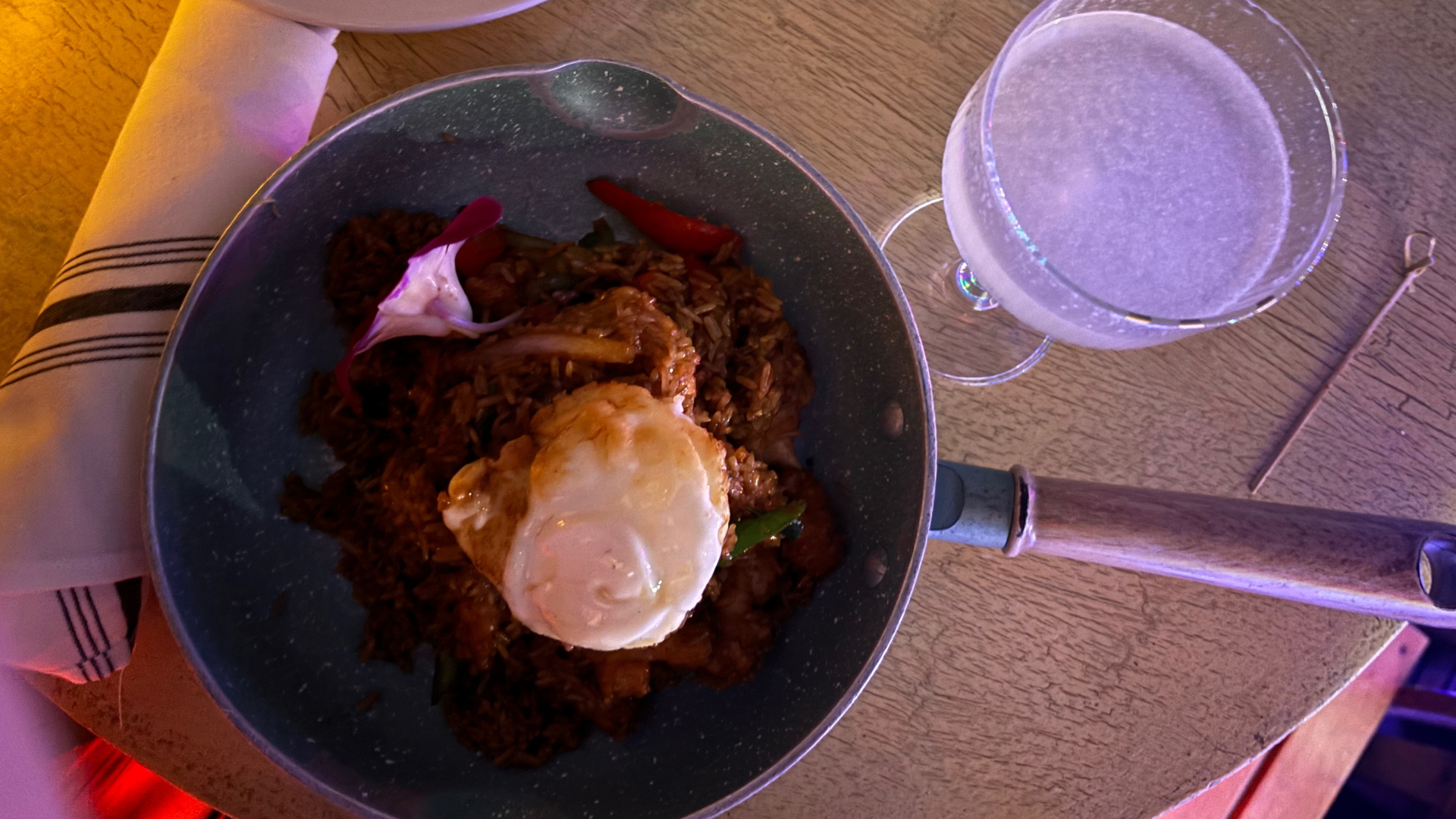 A bowl of rice topped with a fried egg, with some slices of red chili and purple onion, next to a glass of sparkling water on a wooden table, with a napkin and utensils nearby.
