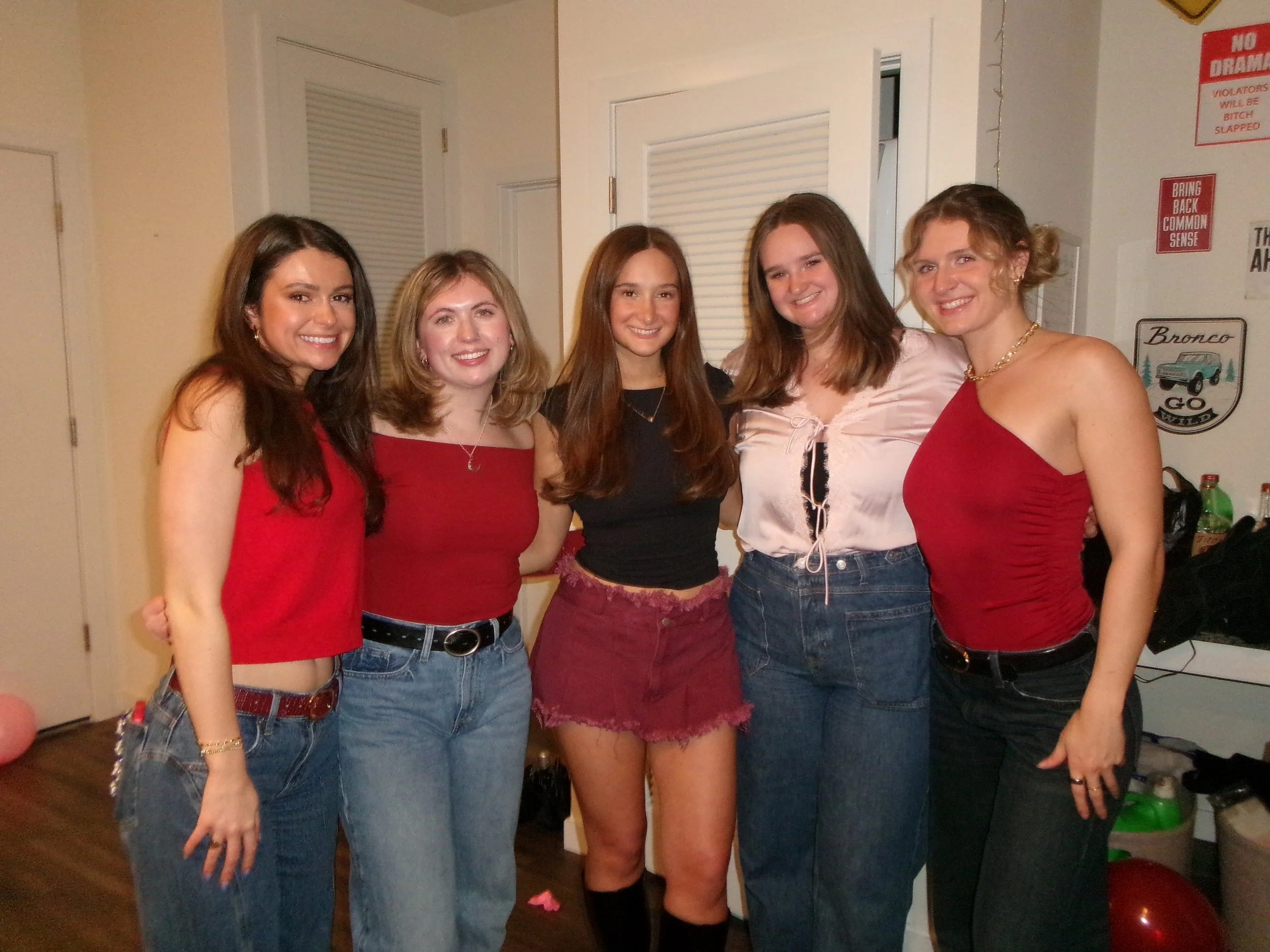Five young women standing together in a room, smiling at the camera. They are dressed casually, with three wearing red tops and two wearing black and white tops.