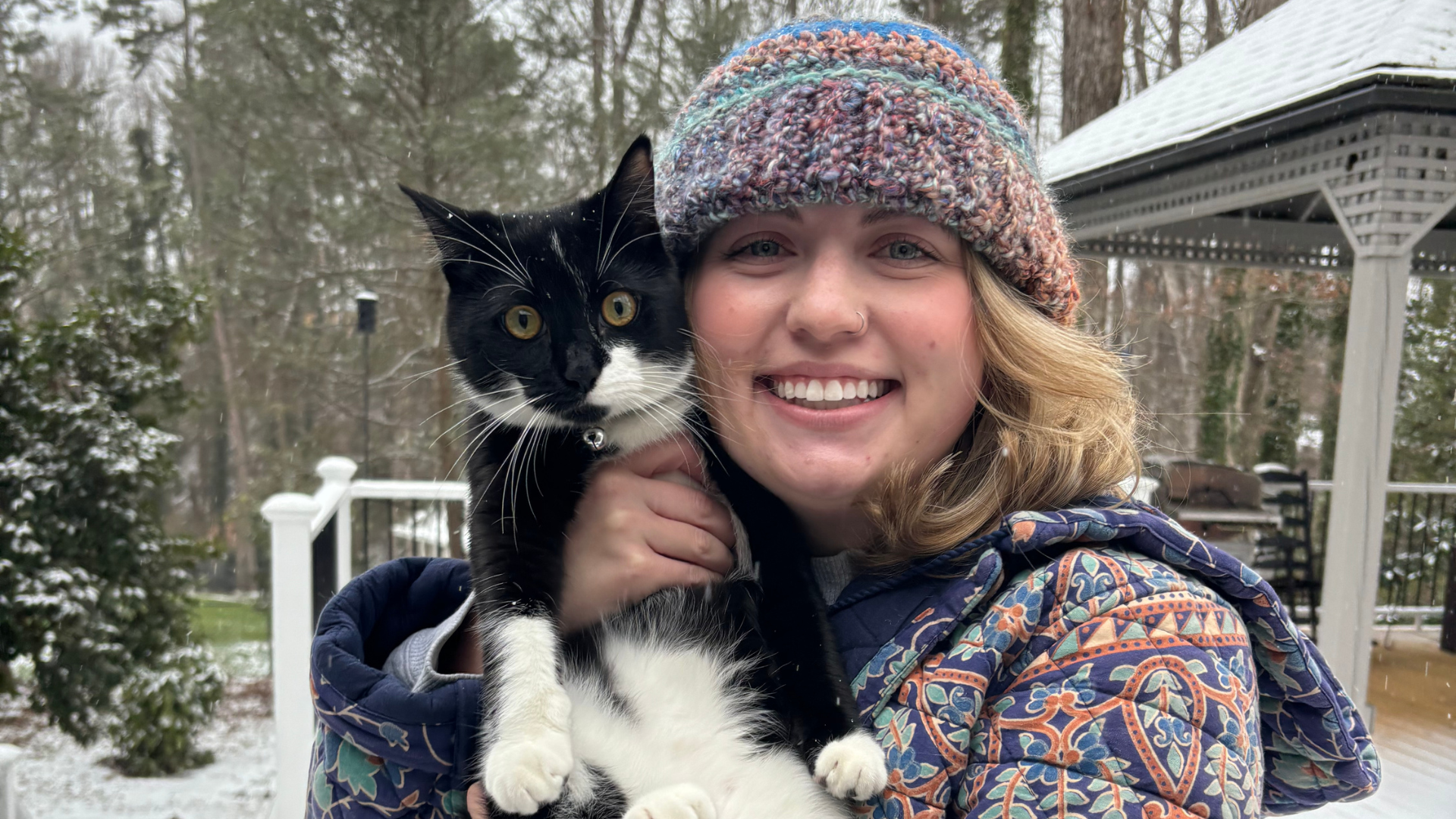 A smiling young woman wearing a multicolored knit cap and a patterned jacket holds a black-and-white cat outside in a winter setting with snow on the roof and trees in the background.