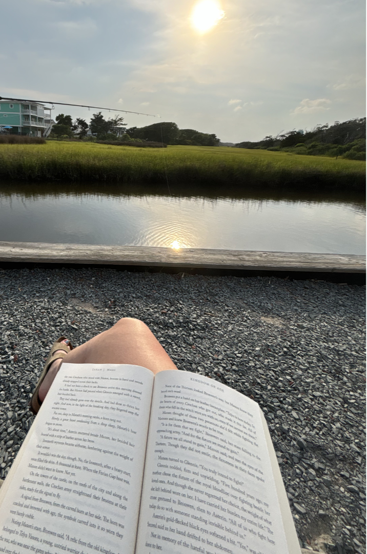 Person relaxing outdoors by a waterway, reading a book, with a grassy landscape, trees, and buildings in the background during sunset.