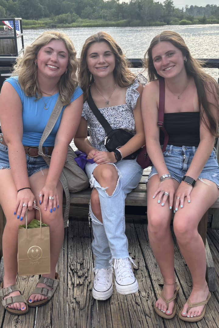 Three young women sitting on a wooden dock by a river, smiling at the camera, with green trees and water in the background.