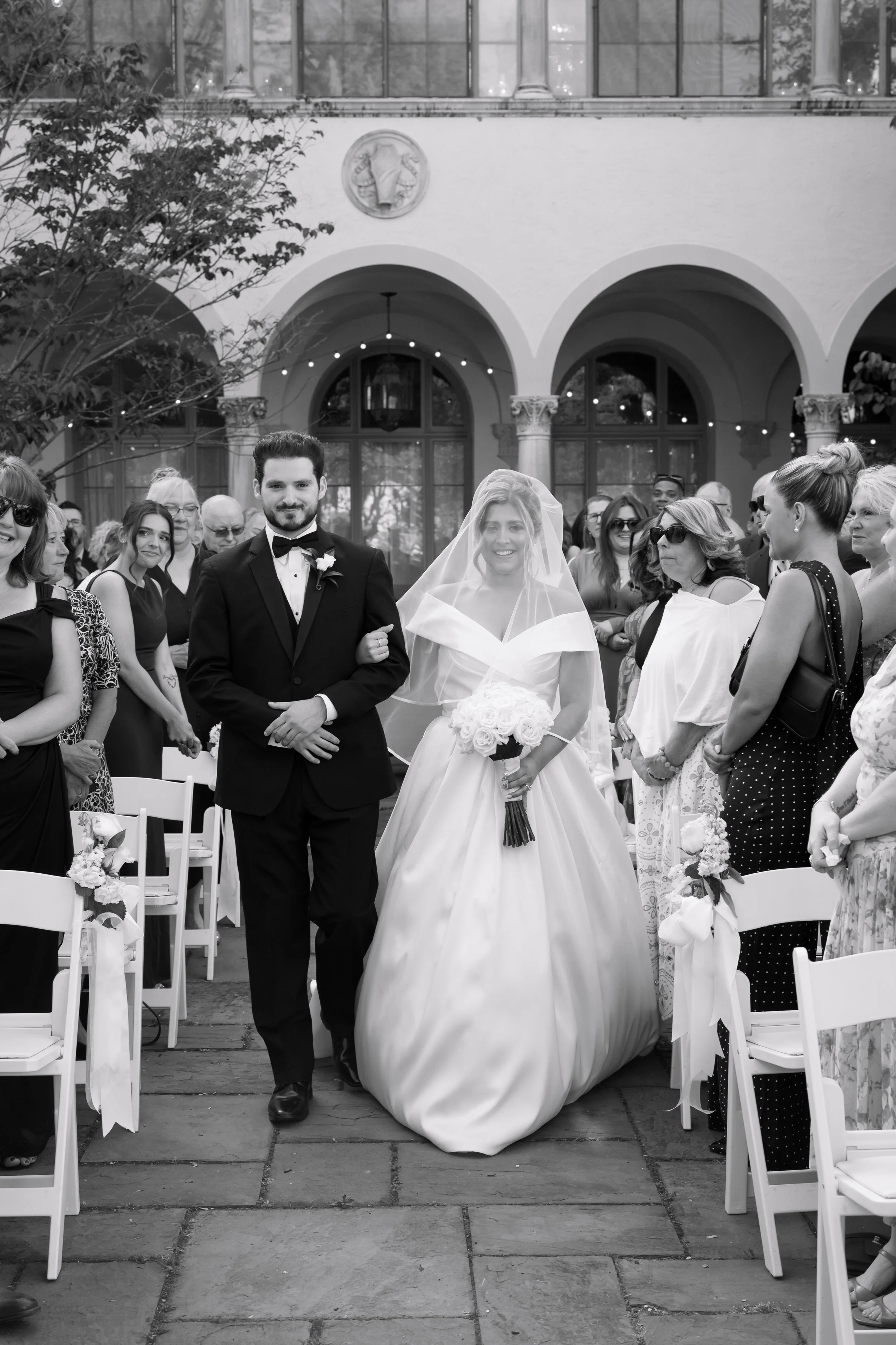A bride and groom share a kiss during their wedding, with the bride in a white dress and the groom in a cream tuxedo holding a small plate.