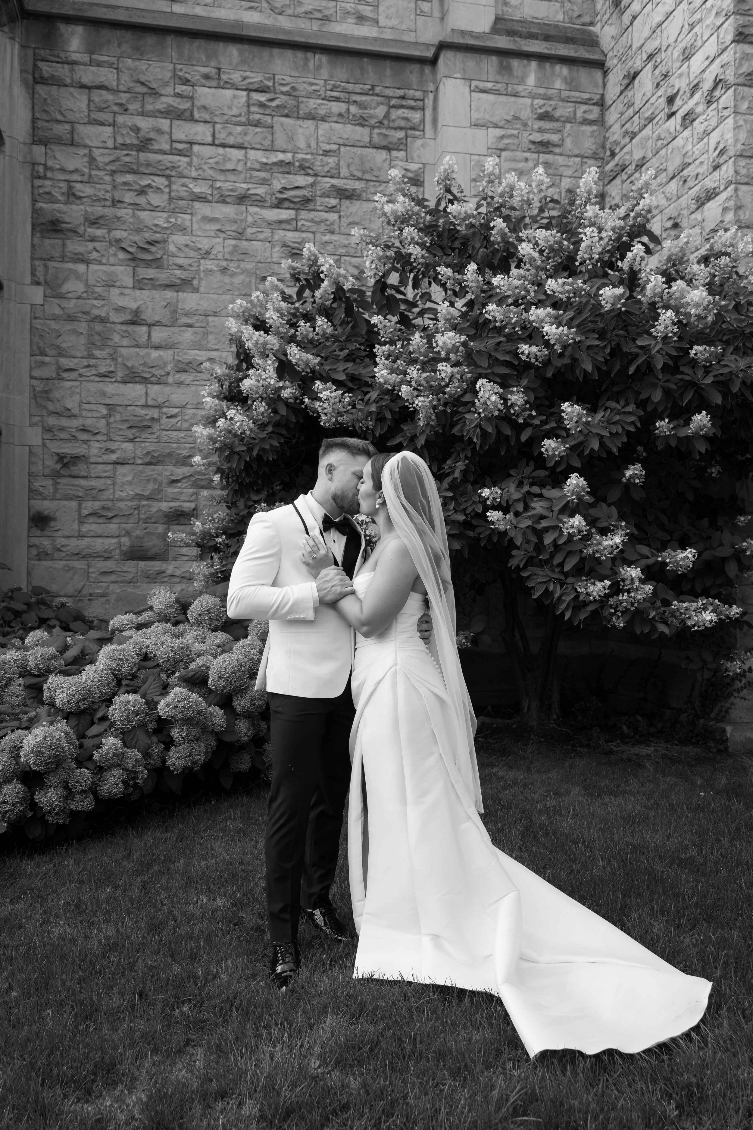 A bride and groom share a kiss during their wedding, with the bride in a white dress and the groom in a cream tuxedo holding a small plate.