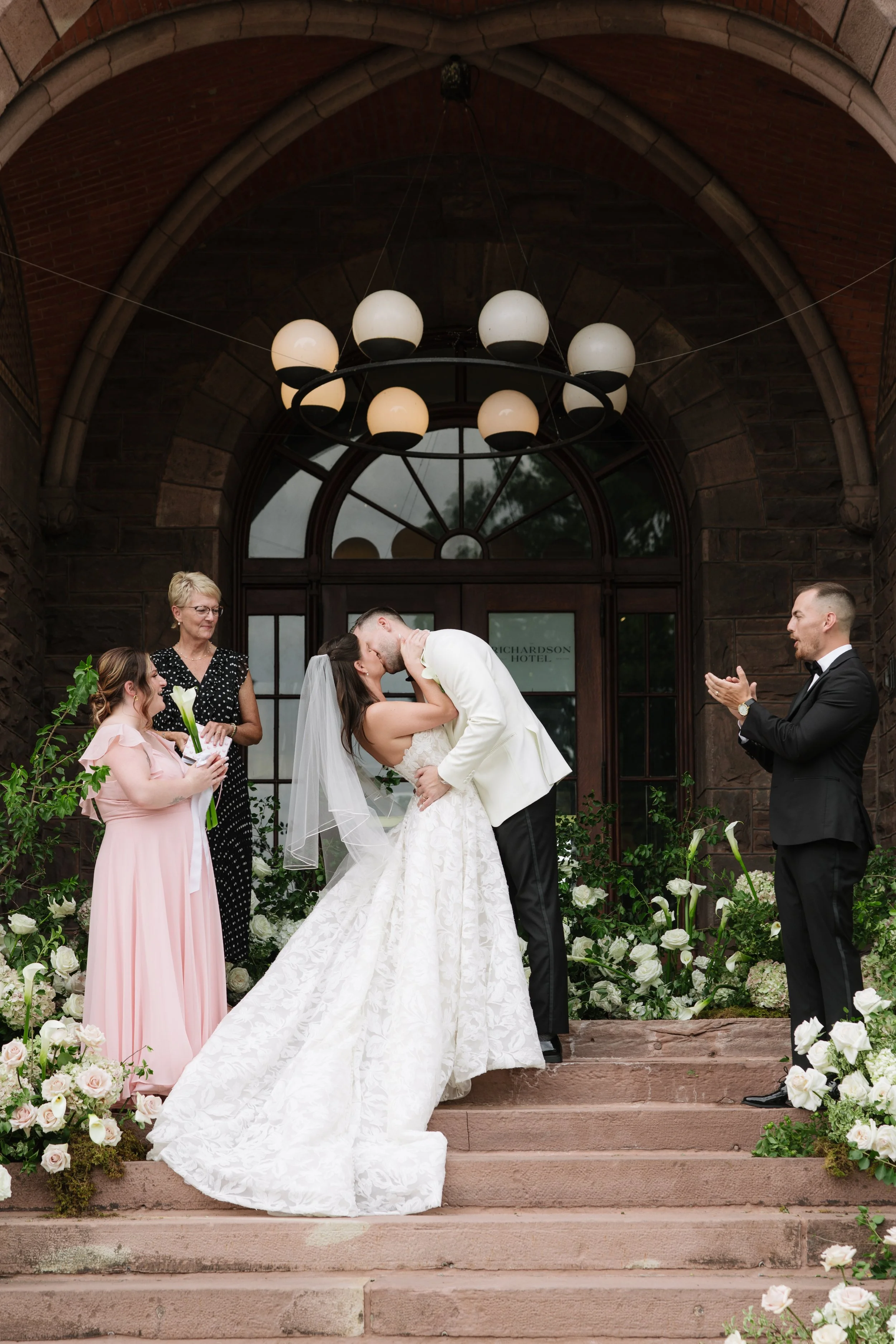 A newlywed couple is kissing outside a grand building with classical columns, while surrounded by friends and family celebrating their wedding, with flower petals being thrown in the air.
