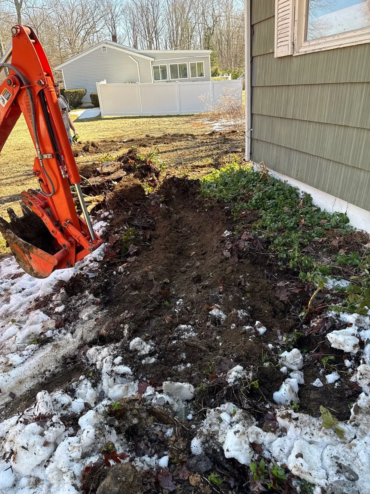 Mini excavator digging a drainage trench beside a residential house with snow and muddy ground in early spring