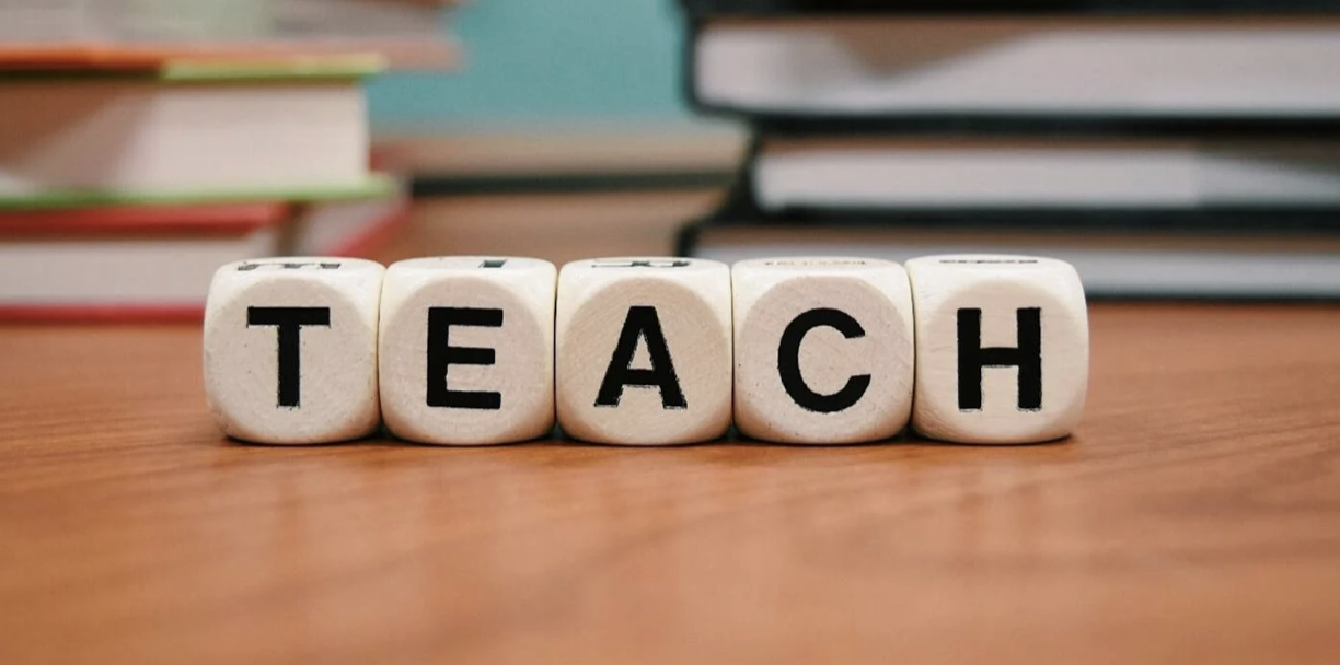 Wooden blocks spelling the word "TEACH" on a table with stacked books in the background.