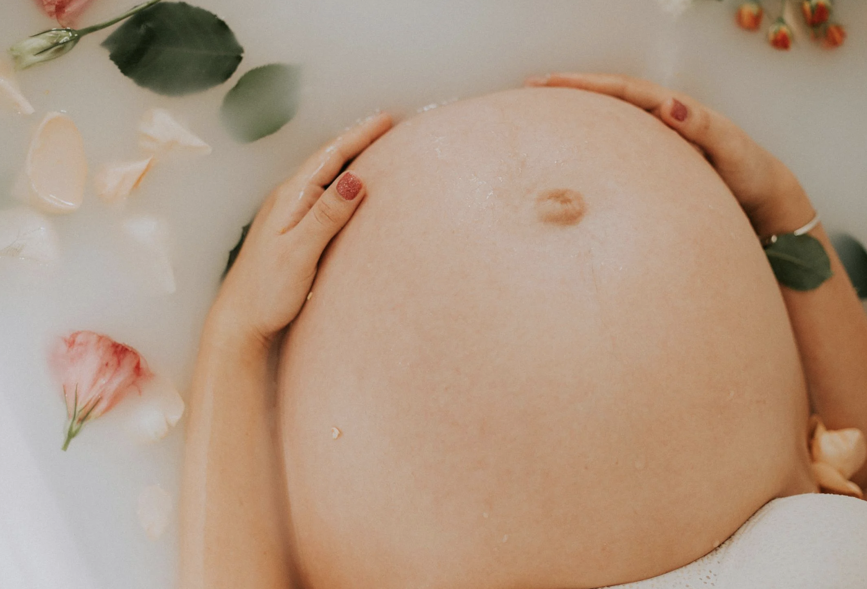 Close-up of a pregnant woman's belly in a bath with floating flower petals and leaves, with her hands gently resting on her belly.