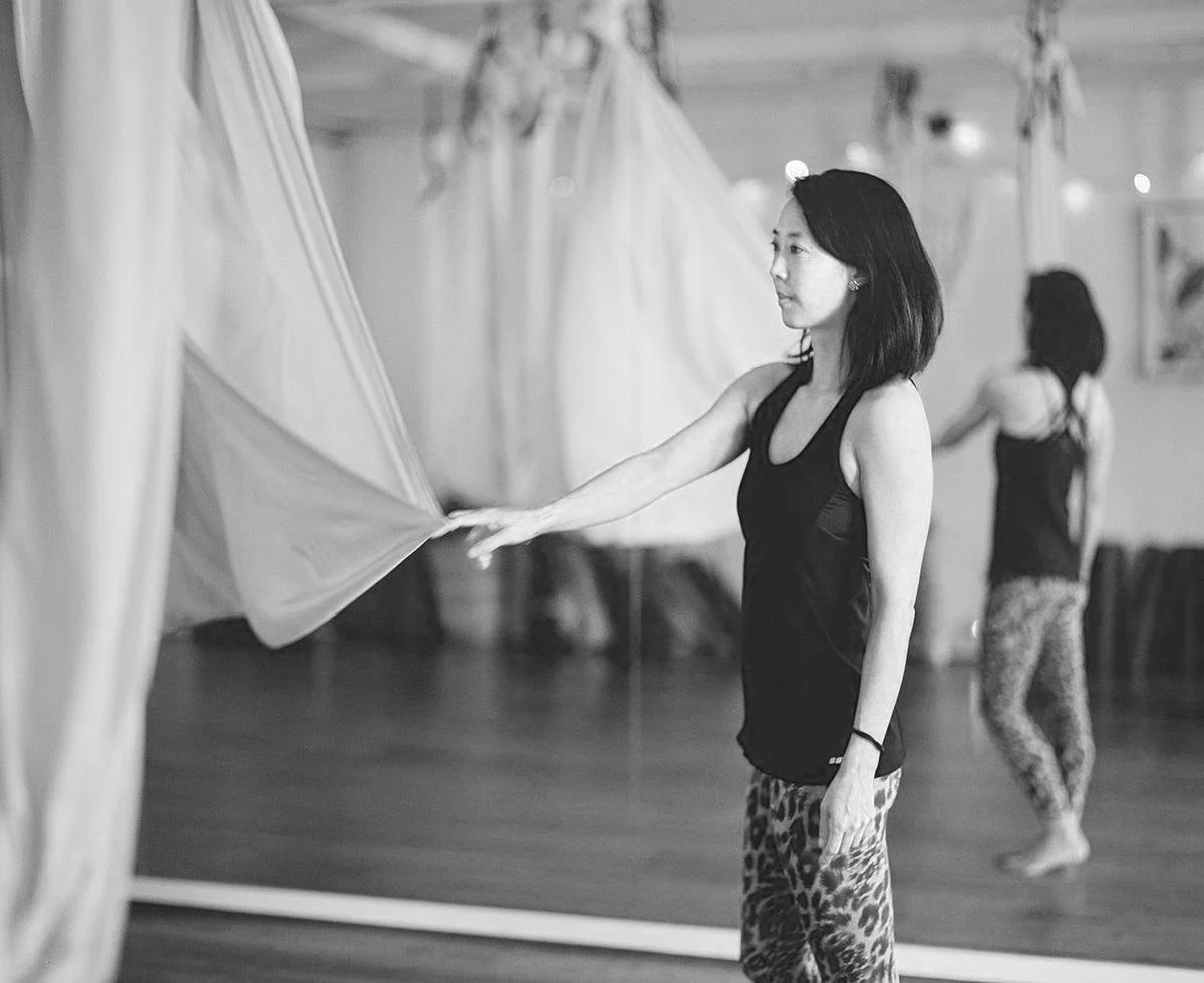 A woman in athletic clothing standing in front of aerial silk in a fitness studio, with a mirrored wall reflecting her image.