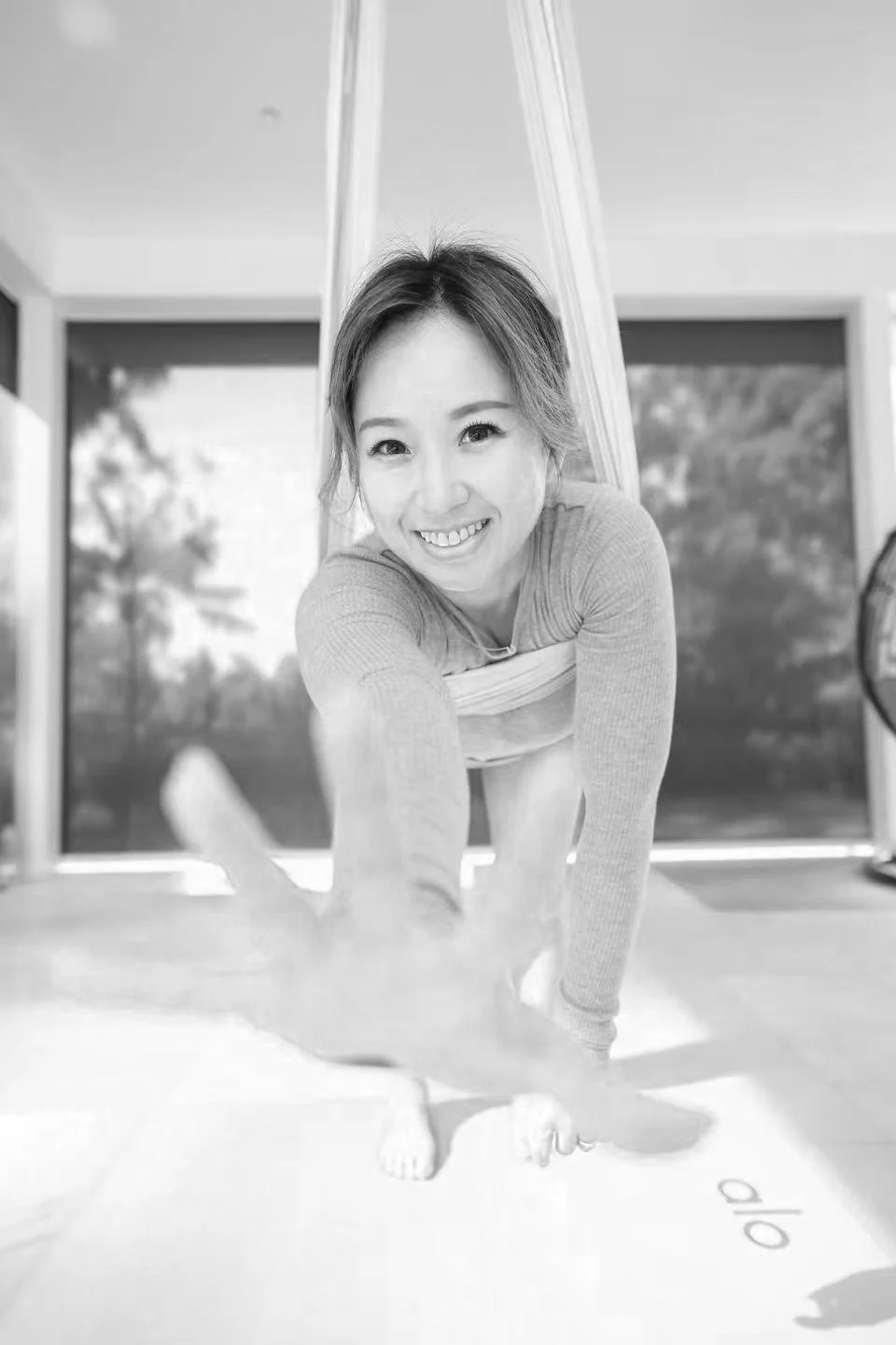 Woman practicing aerial yoga indoors, smiling and reaching out towards camera, black and white photo.