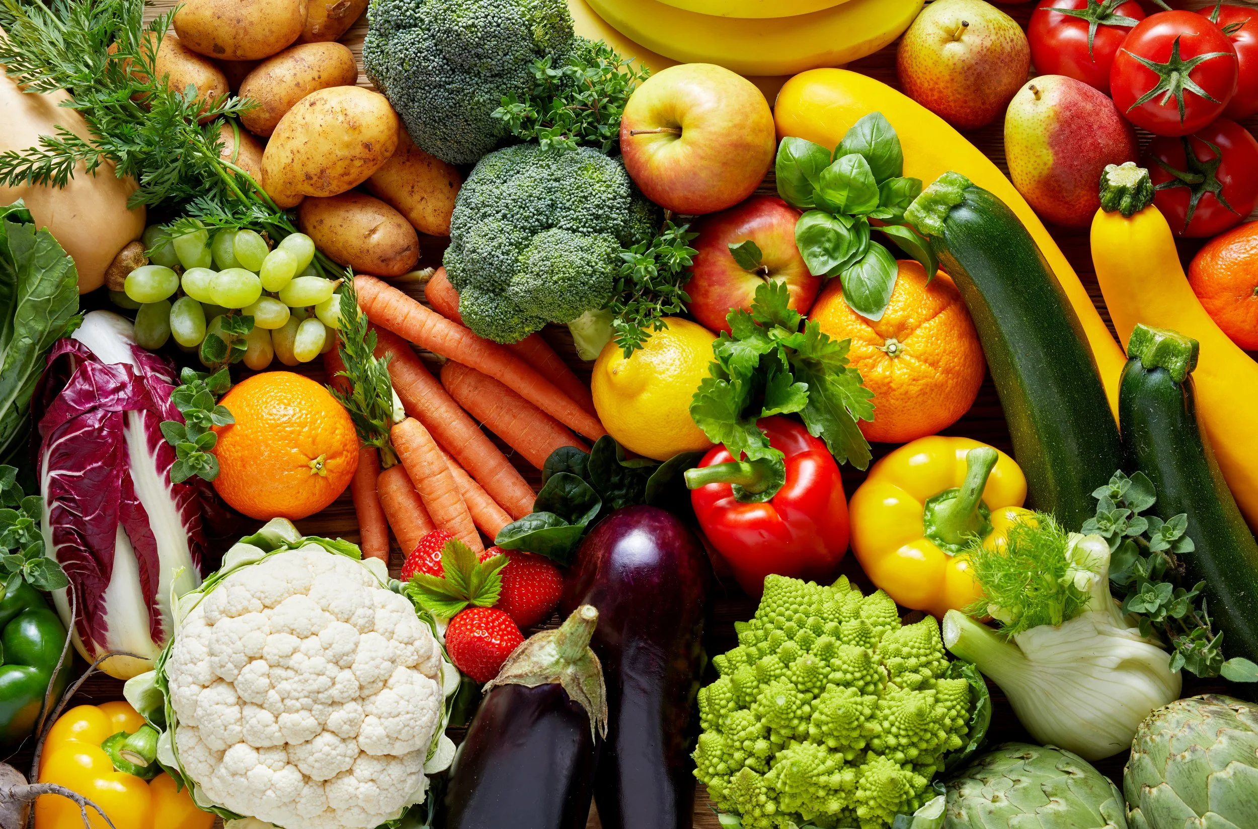 A colorful assortment of fresh fruits and vegetables including tomatoes, zucchini, carrots, grapes, bell peppers, cauliflower, broccoli, eggplant, strawberries, and oranges.