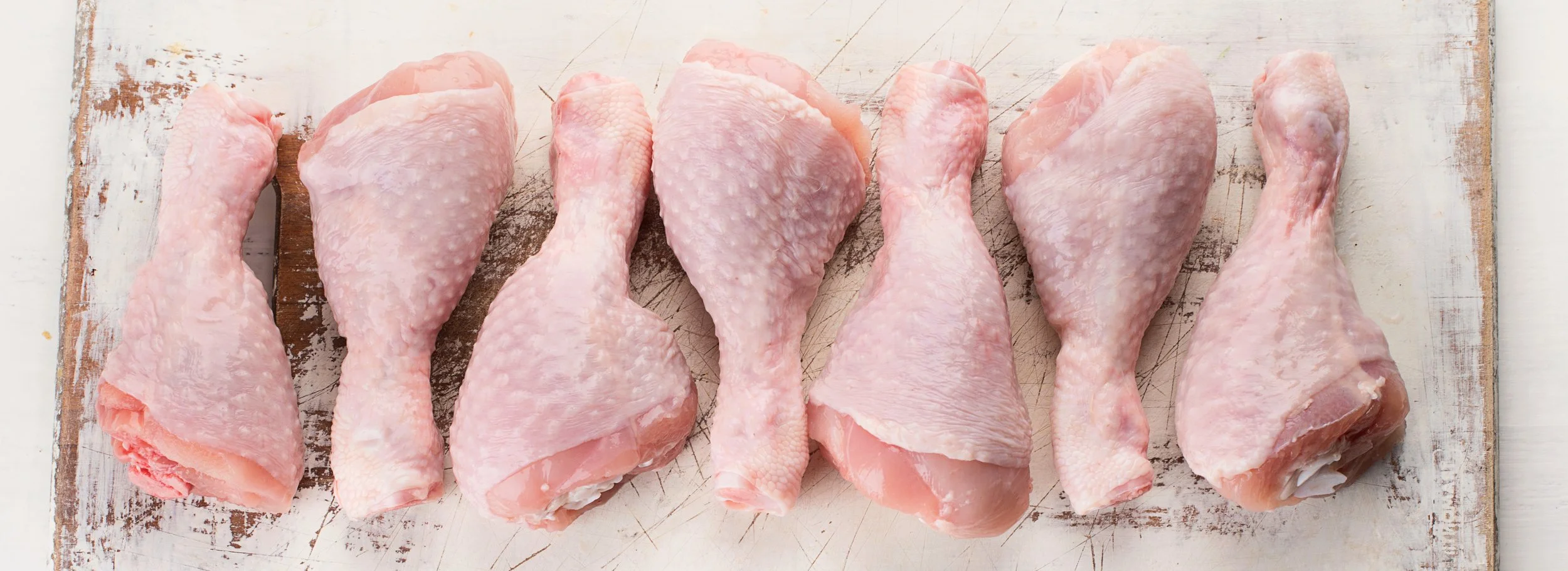 Seven raw chicken drumsticks arranged on a rustic white wooden cutting board.