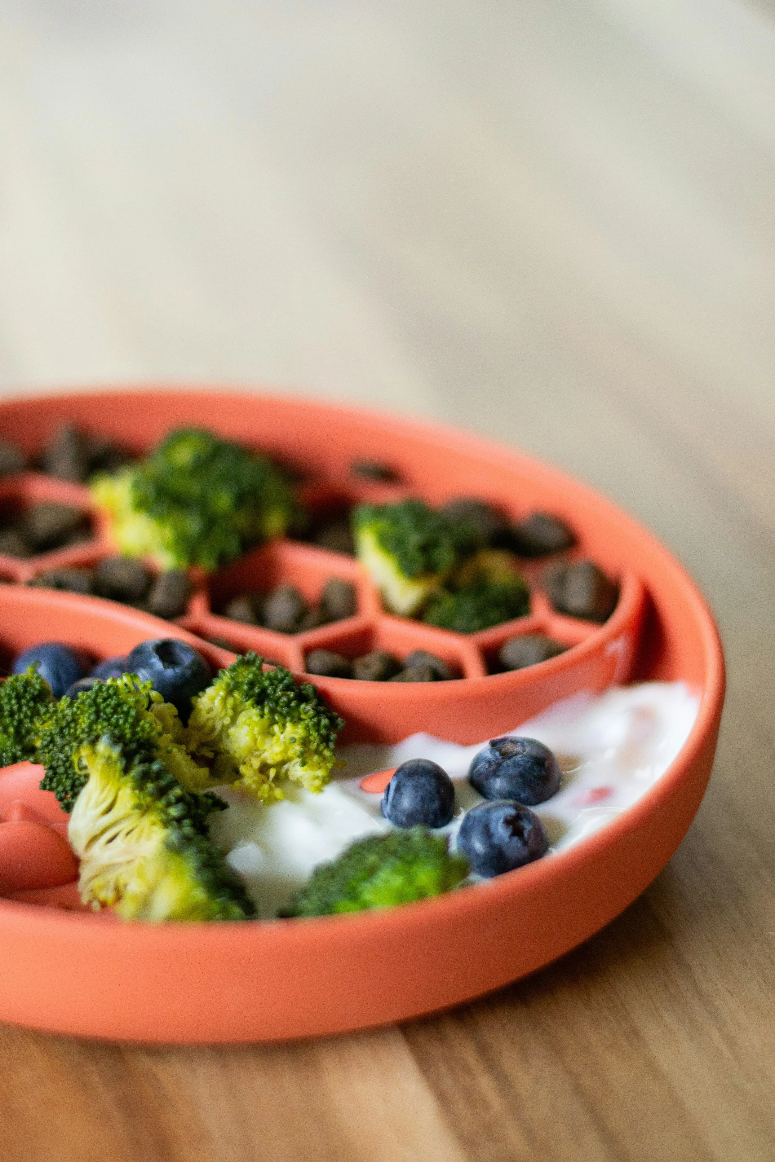 A pink divided snack tray filled with broccoli, blueberries, and a yogurt dip, placed on a wooden surface. An example of fresh toppers to enhance a commercial diet.