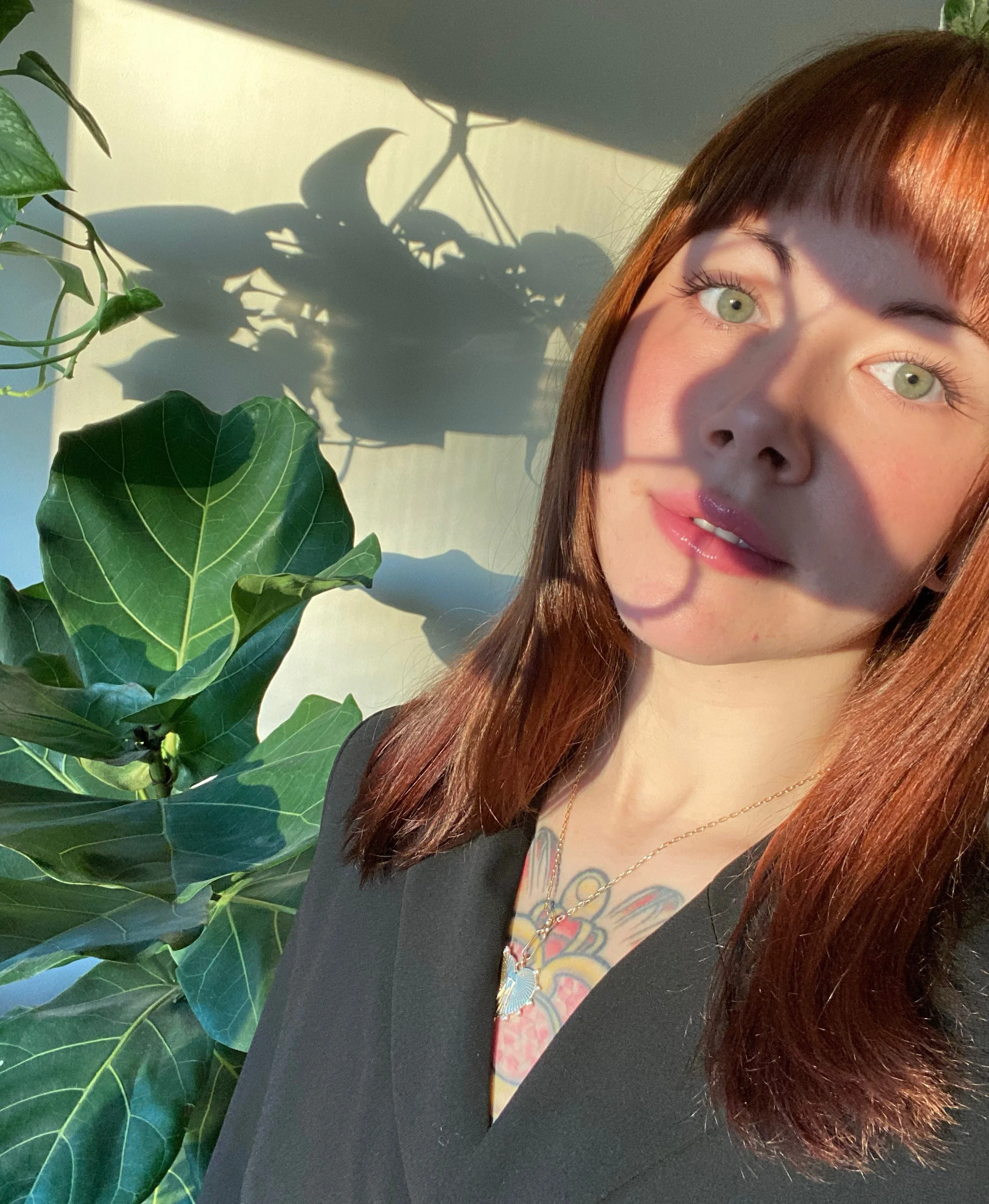 A young woman with reddish-brown hair and green eyes taking a selfie next to a large green plant, with sunlight creating shadows on the wall behind her.