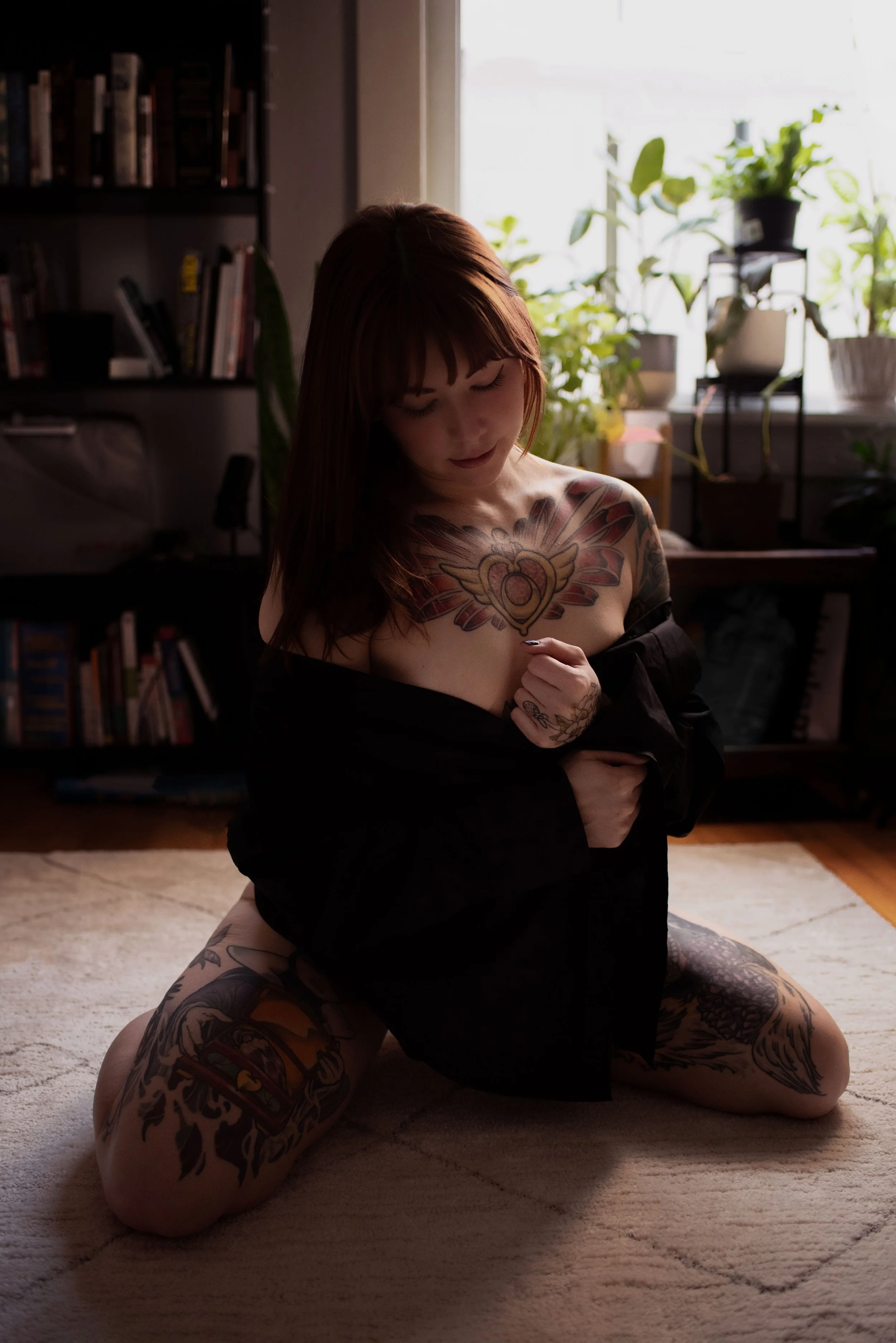 A woman with tattoos kneeling on a beige carpet in a room with a bookshelf and potted plants near a window. She is partially covered with a black shirt, looking down with her eyes closed.