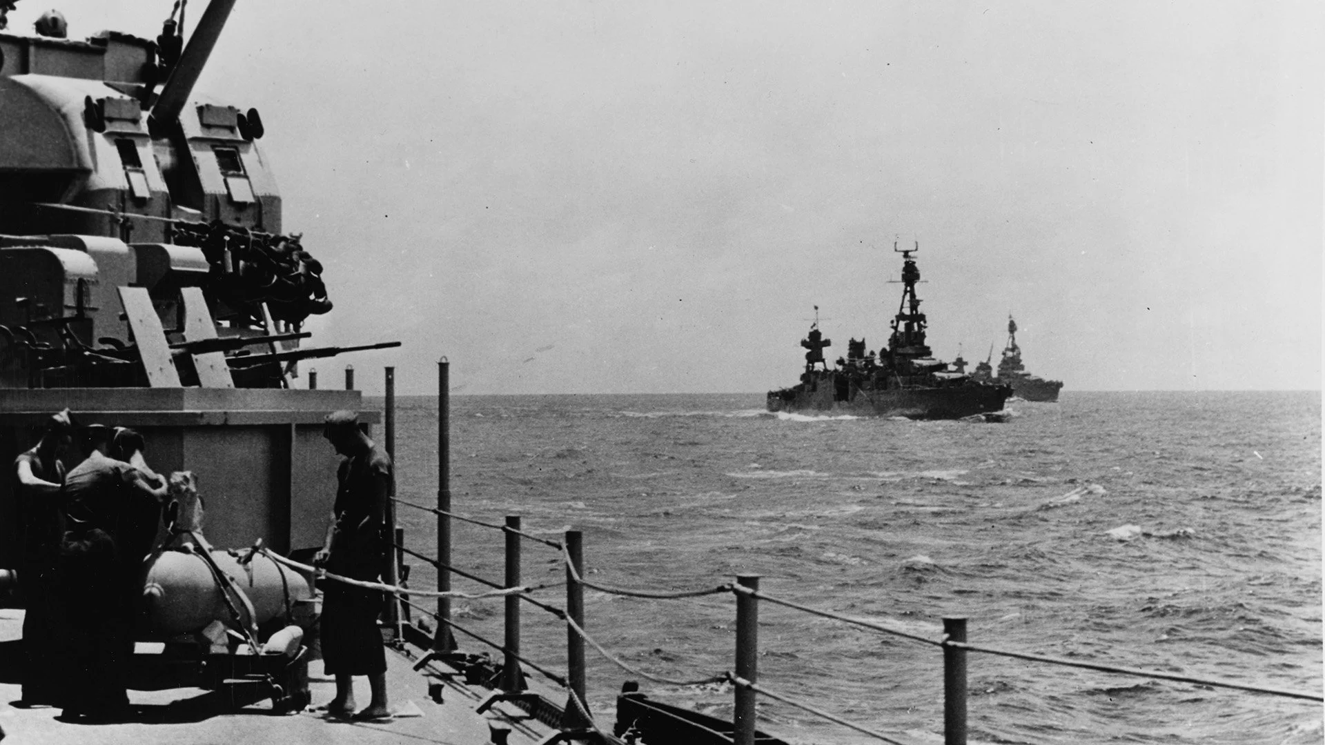 Black and white photo of naval ships sailing on the ocean, with crew members working on the deck in the foreground.