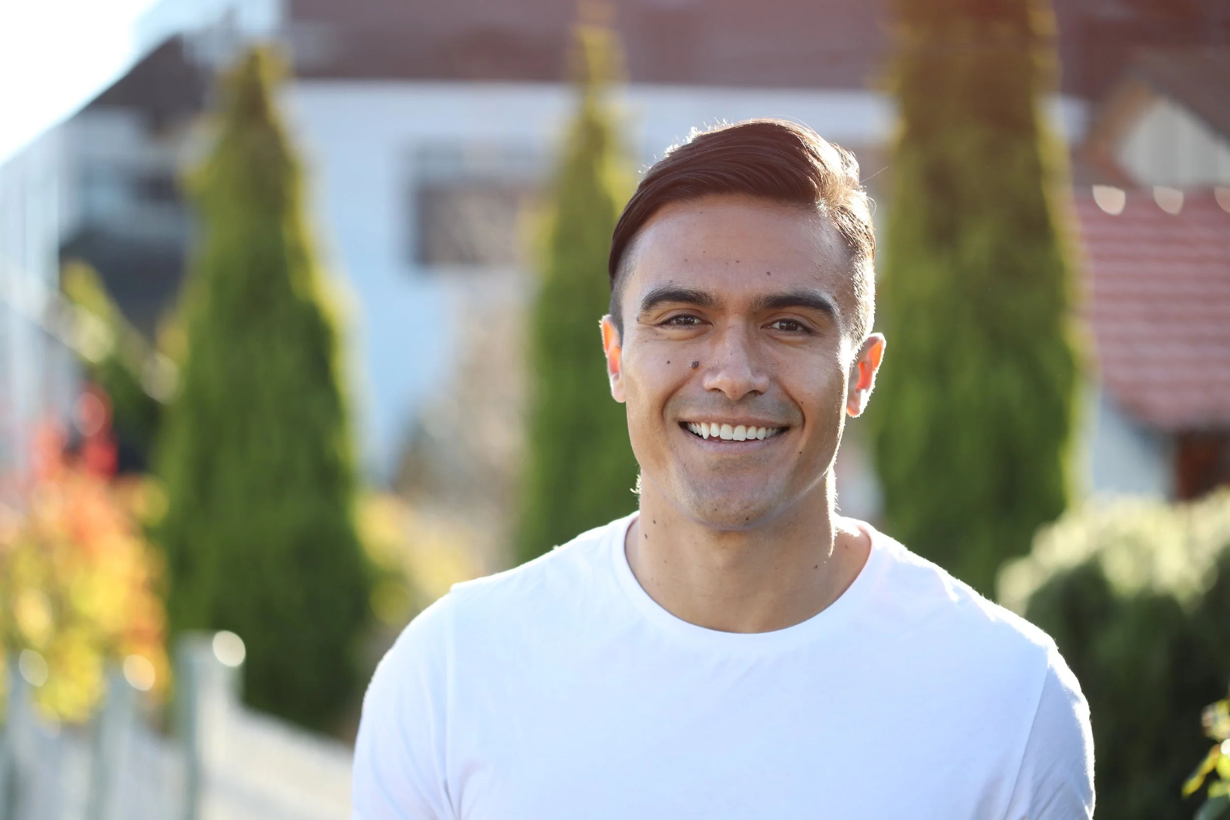 A young man with short brown hair smiling outdoors on a sunny day, wearing a white t-shirt, with green trees and houses in the background.