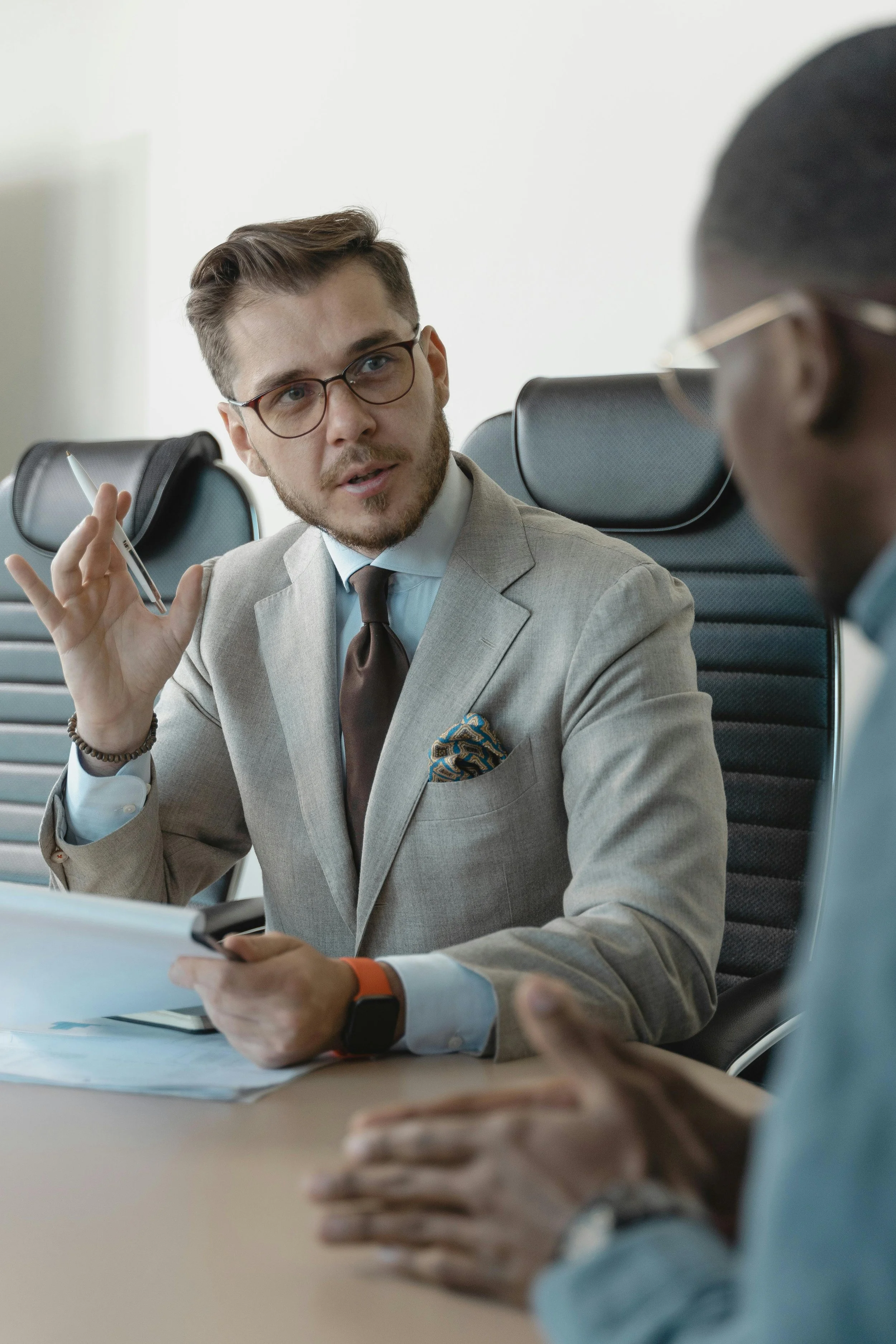 Two men having a business meeting in a modern office, one is speaking and holding a pen, the other is listening and resting his hands on the table.