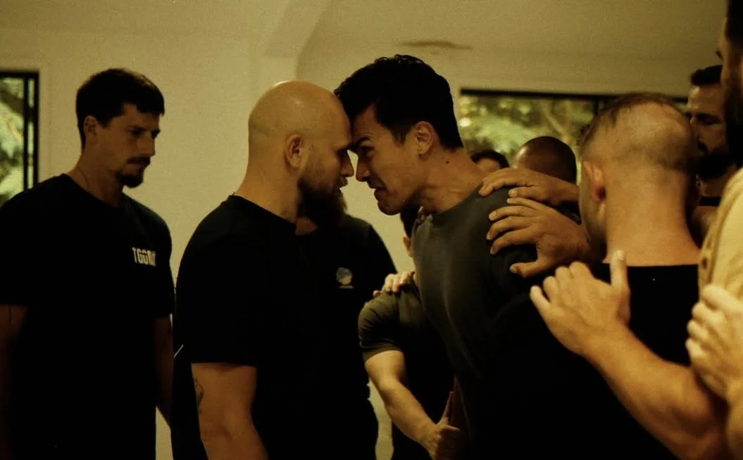 Two men, with one bald and bearded and the other with dark hair, face off in a tense stare-down while being held back by other people, in what appears to be an intense confrontation.