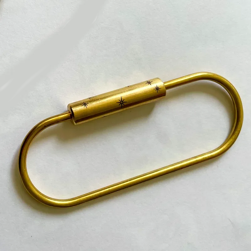 Close-up angled view of two brass oval carabiner key rings with screw-lock closures engraved with starburst star details. Matte brass finish shown in natural light on white background.