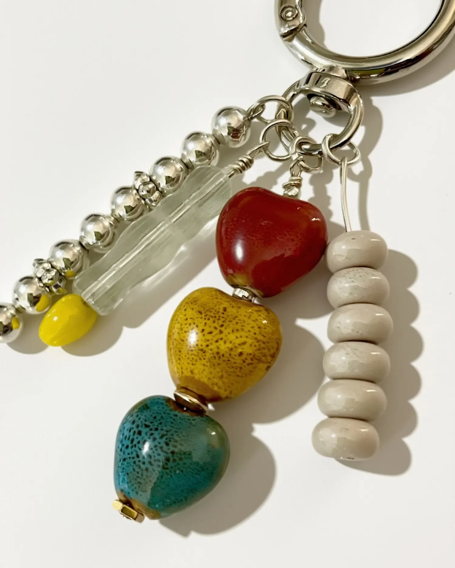 Silver carabiner keychain with red, mustard, and teal ceramic heart beads, a clear glass bead, yellow accent bead, and white ceramic rondelle beads on white background.