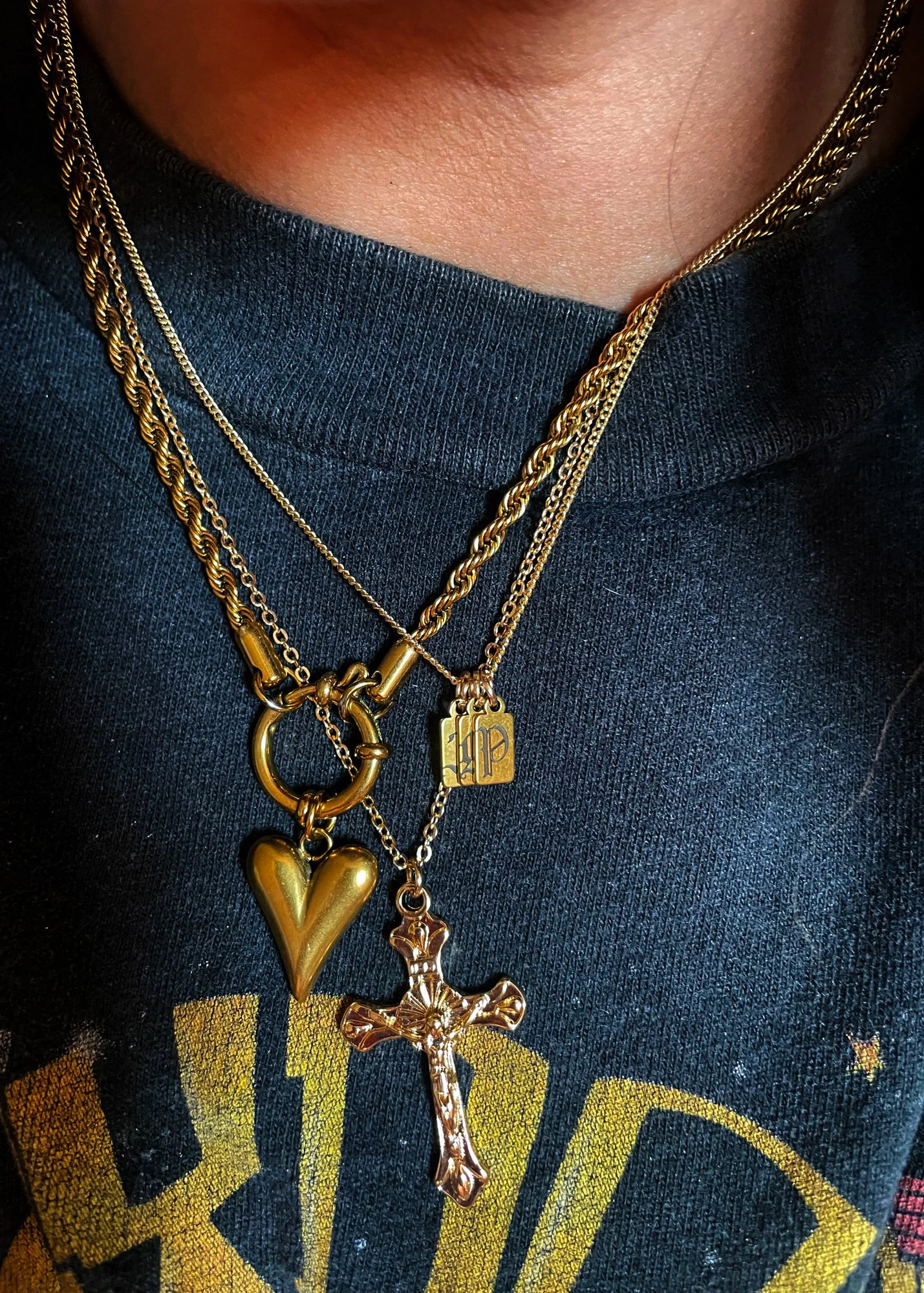 Multiple layered gold necklaces with pendants, including a cross, a heart, and a tag with initials, worn over a dark shirt.