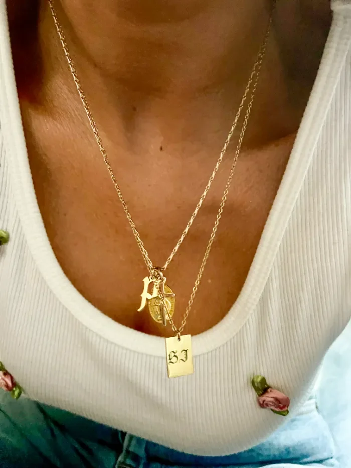 women wearing white tank top with tiny pink silk roses on it wearing two gold necklaces layered over each other, one of them engraved in old english gothic font "SJ" and a blackletter "p" pendant, mini gold cross charm and mini Gold Miraculous Medal