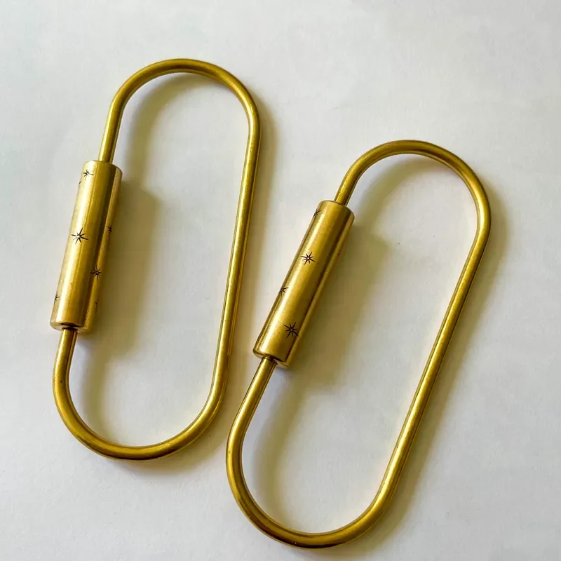 Two solid brass oval screw-lock carabiner key rings with engraved starburst barrel details laid flat on a white surface. Warm matte gold finish. Just under 3.5 inches in length.