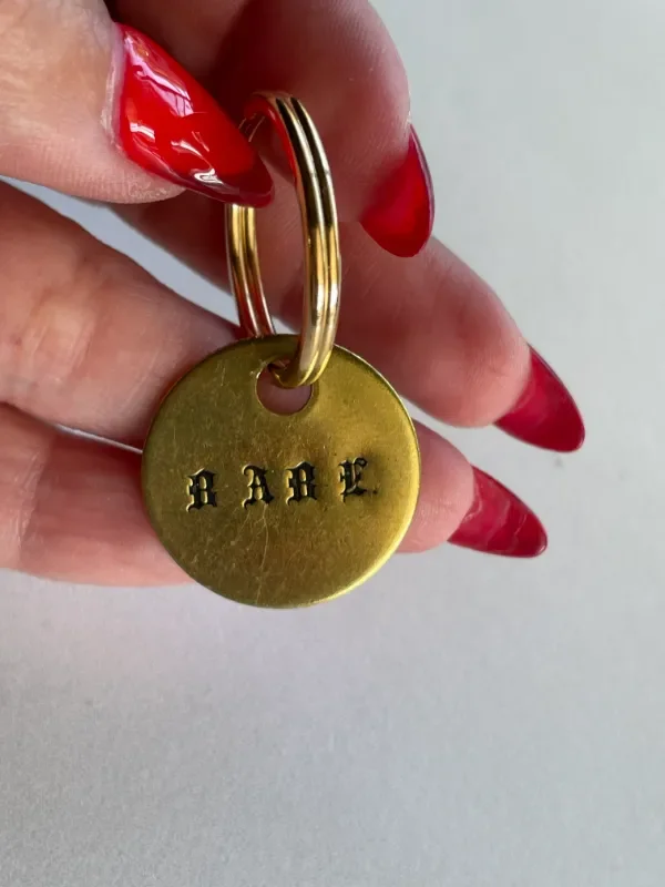and holding a brass keychain tag stamped with "BABE" in Gothic blackletter font with a gold split ring