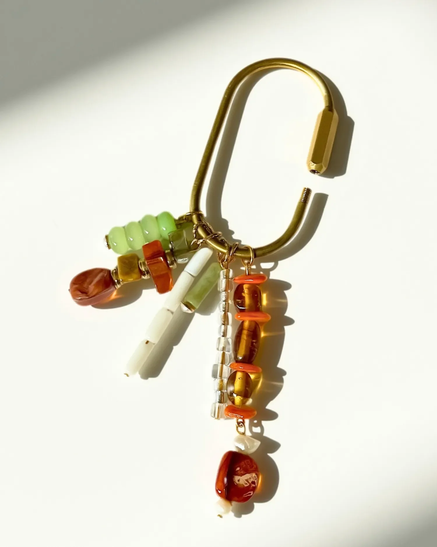 Brass carabiner charm shown open, with the beaded strands — amber, coral, mint, and pearl — fanned out to the side.