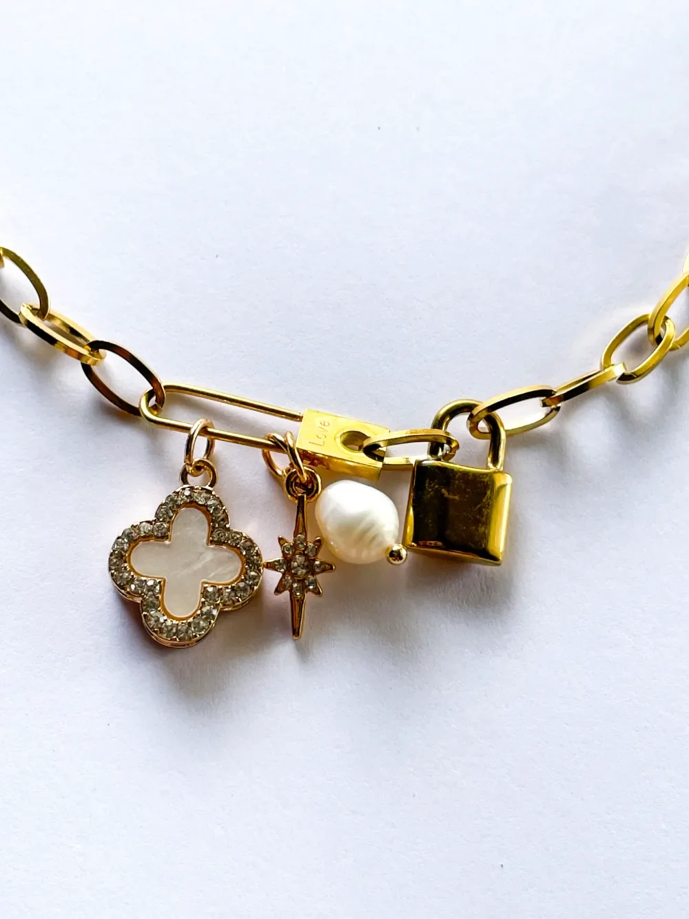 Close-up of gold charm necklace featuring pavé clover, freshwater pearl, starburst, and gold locket engraved Love on paperclip chain