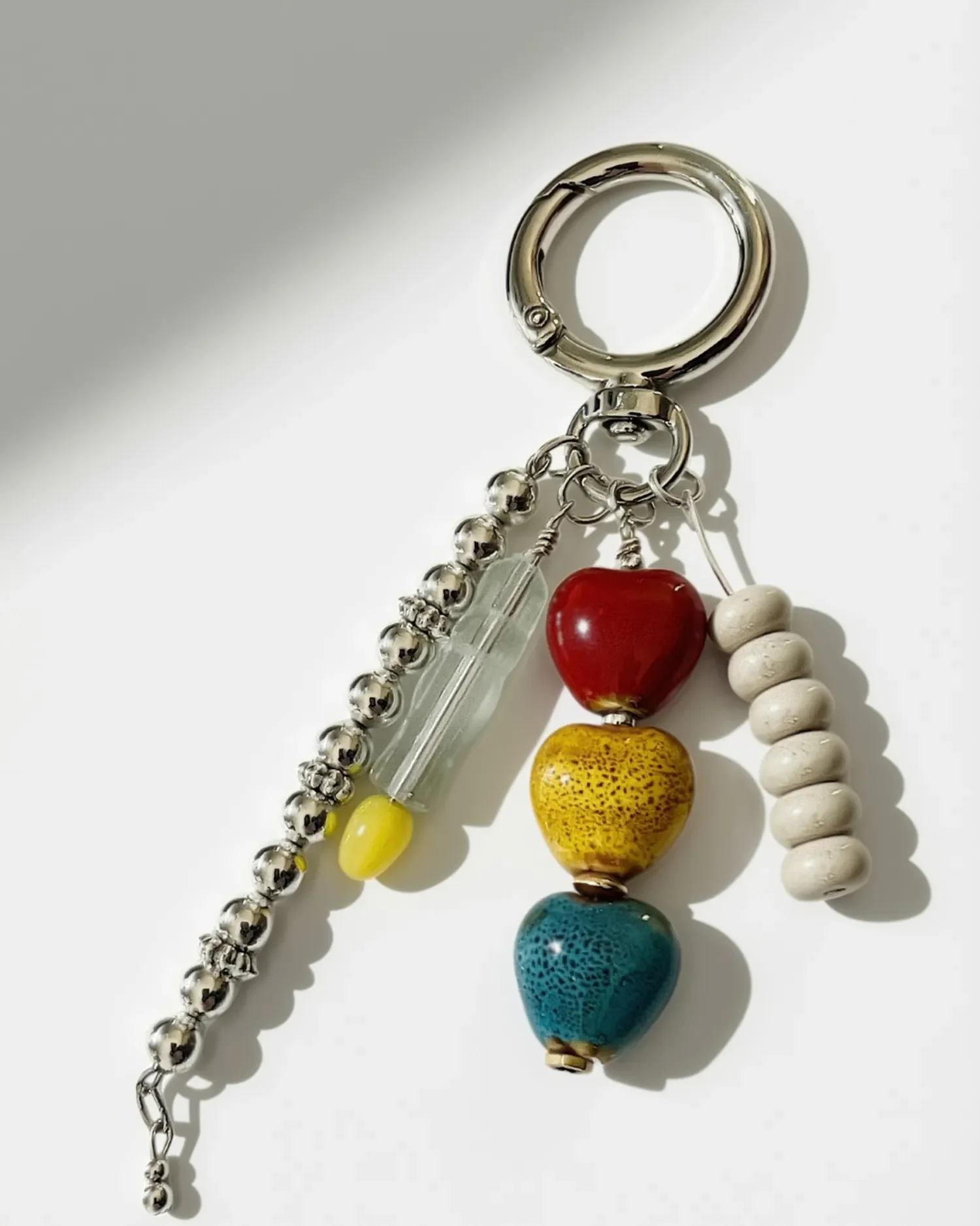 Close-up of a beaded bag charm: red, mustard, and teal ceramic heart beads next to a clear glass bead with yellow accent and a stack of cream ceramic rondelle beads.