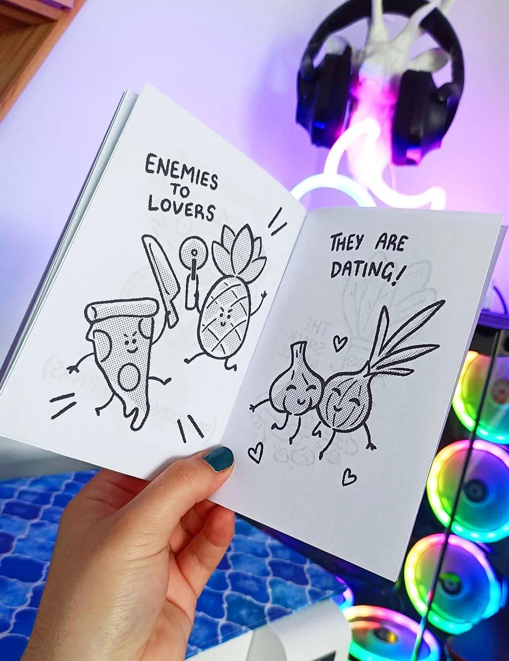 Open sketchbook with cartoon drawings of food items, fish, and vegetables with humorous text about enemies and lovers, and a playful dating message, held in front of a neon-lit background with colorful lights.