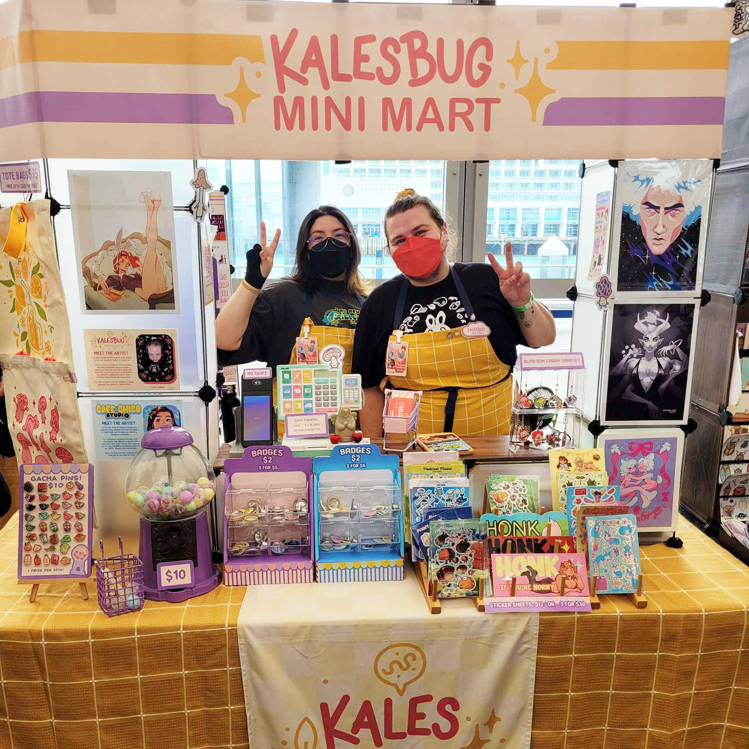 A booth at a convention or market called 'Kalesbug Mini Mart' decorated with pastel-colored signs, featuring various small items including stickers, magnets, pins, and character artwork. Two people wearing masks are standing behind the booth, making peace signs.