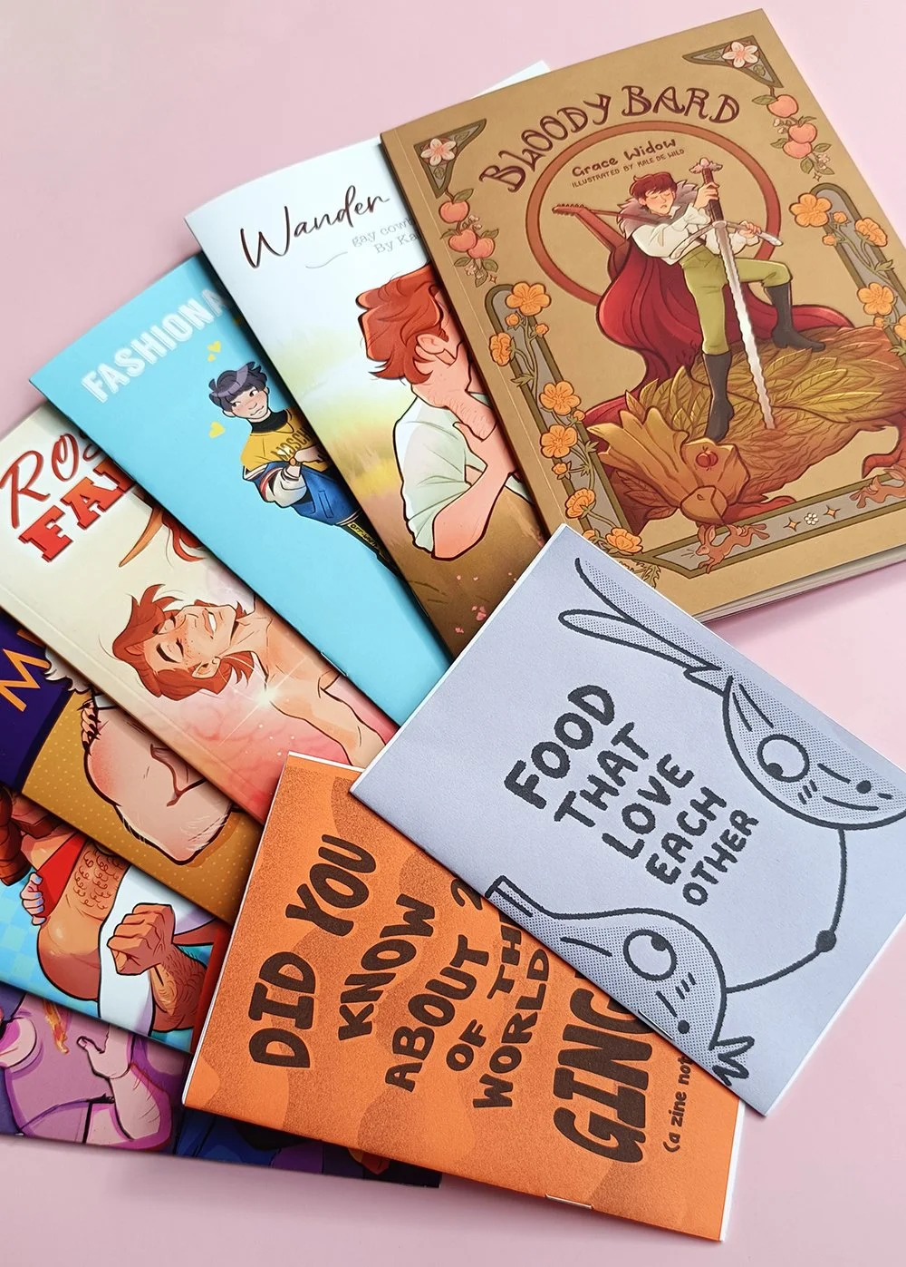 A collection of zines with colorful covers, including titles like "Bloody Bard" and "Food That Love Each Other," arranged on a pink surface.