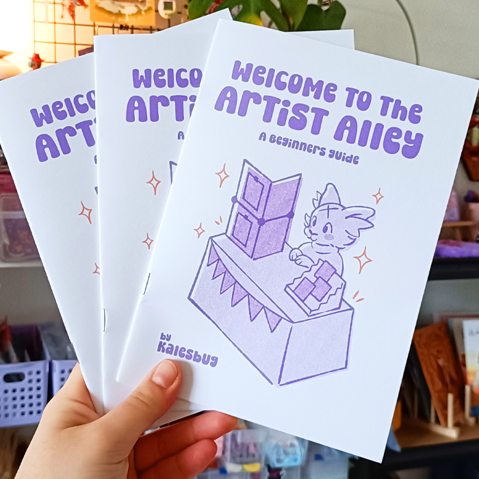 Three pamphlets titled "Welcome to the Artist Alley: A Beginner's Guide" held in a person's hand, with a cartoon illustration of a cat at a table with a geometric sculpture, in a colorful indoor setting.