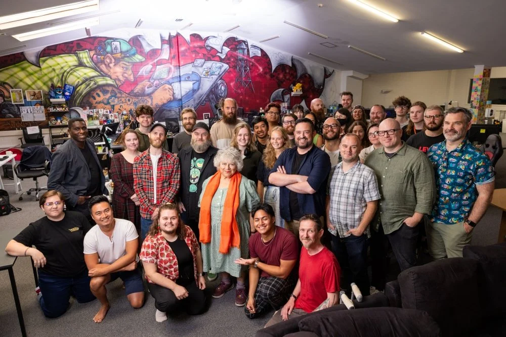 Group of diverse people smiling and posing for a photo in an office with a large graffiti mural of a person in a hat and a dog on the wall.