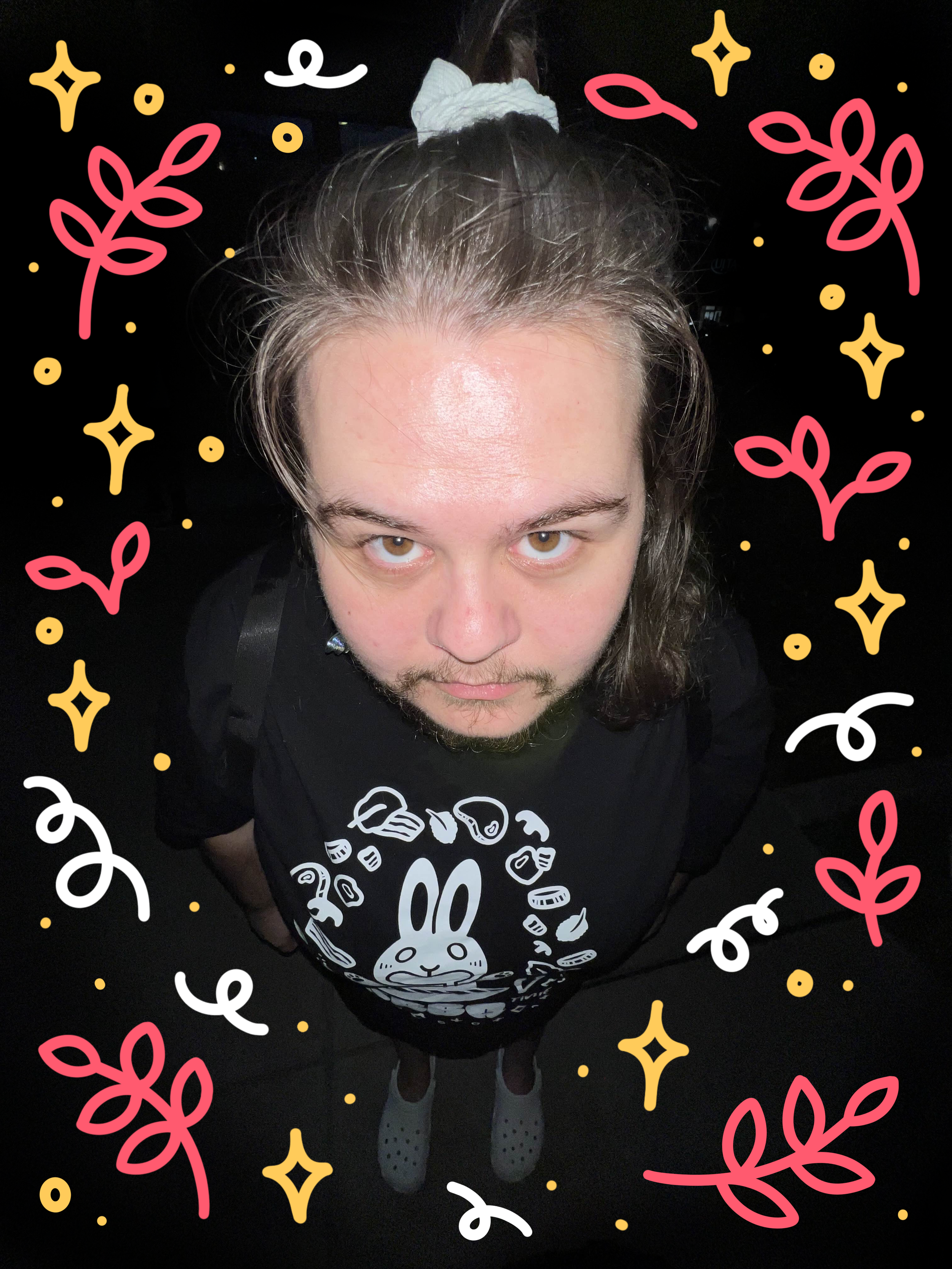 A person with brown hair tied up in a white scrunchie, wearing a black t-shirt with a white bunny design and food illustrations, standing against a dark background with colorful decorative elements around the border.