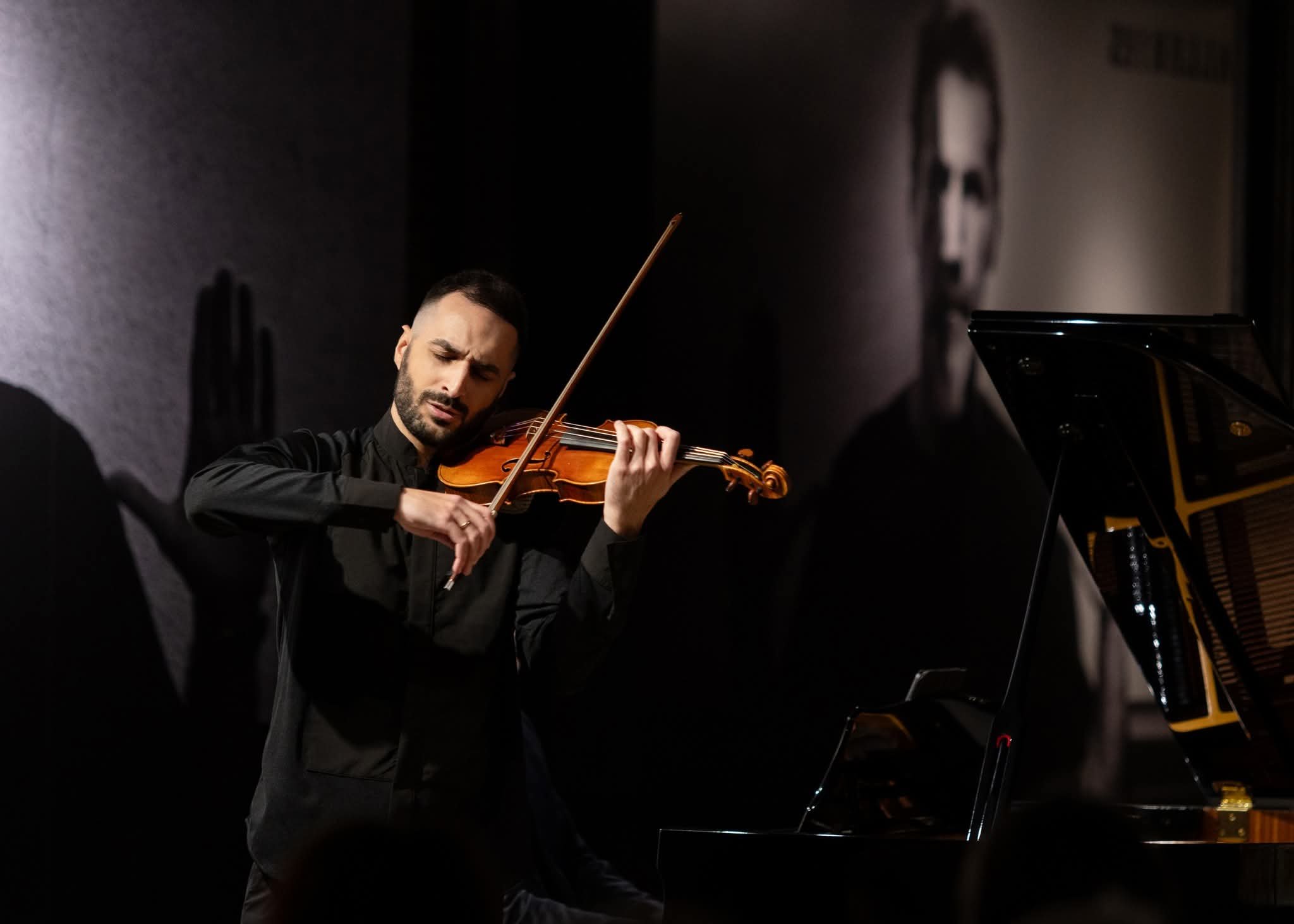 A man playing a violin on stage, dressed in black, with a grand piano nearby.