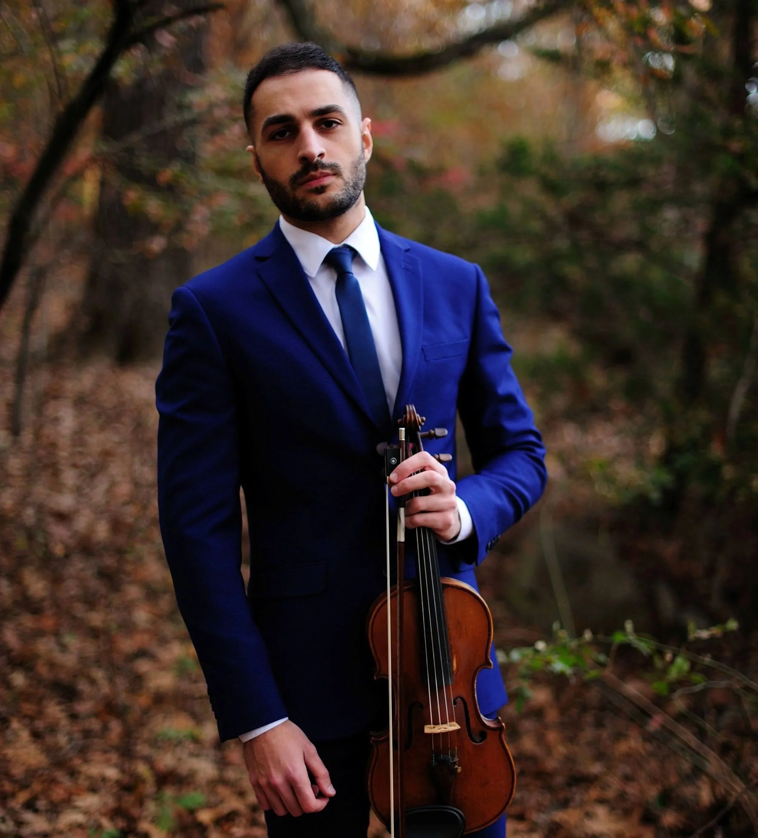 A man in a blue suit and white shirt holding a violin and bow, standing outdoors in a wooded area with fall foliage.