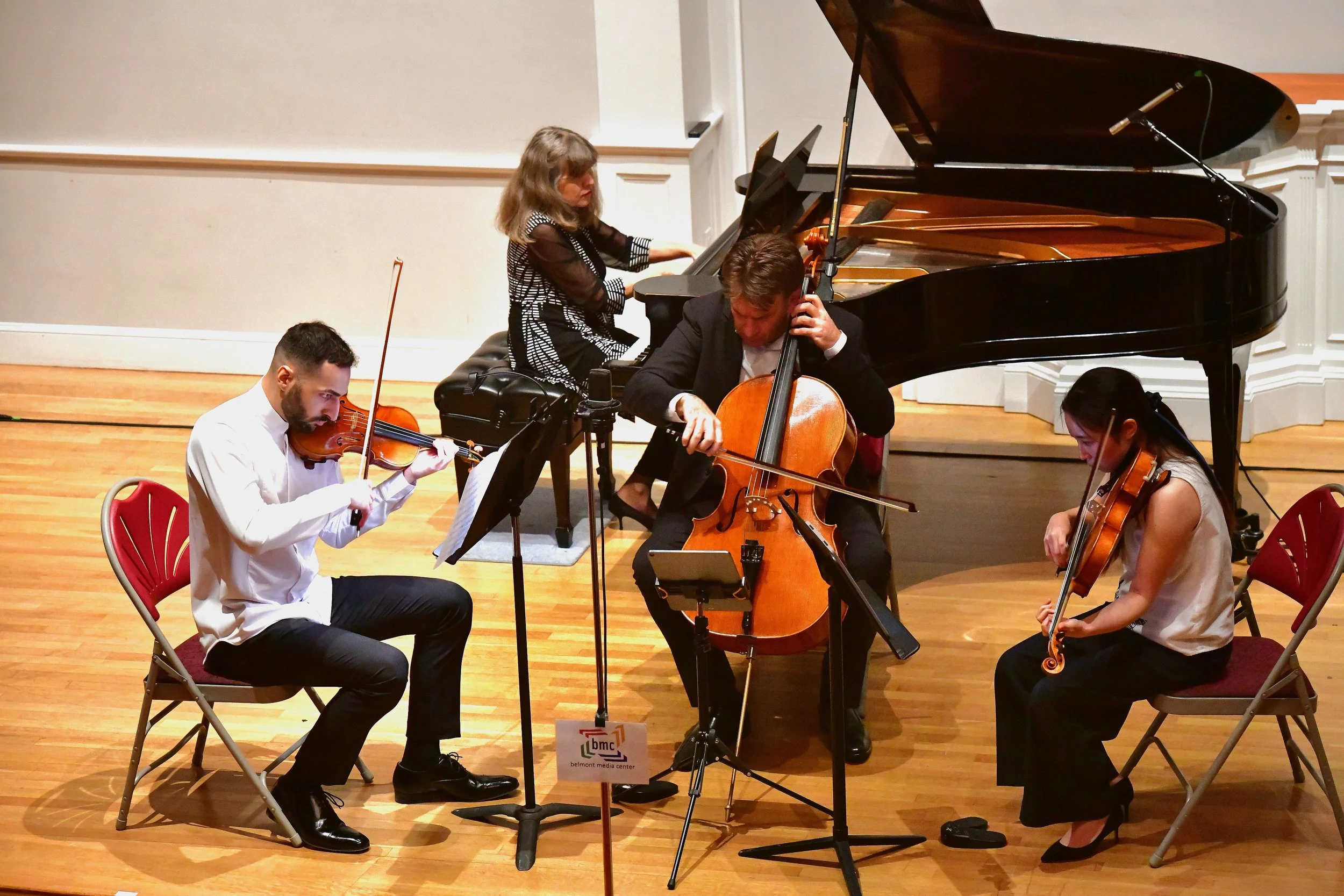 Four musicians performing on stage with a grand piano: a violinist, a cellist, a pianist, and a violist, all focused on their music in a concert hall.