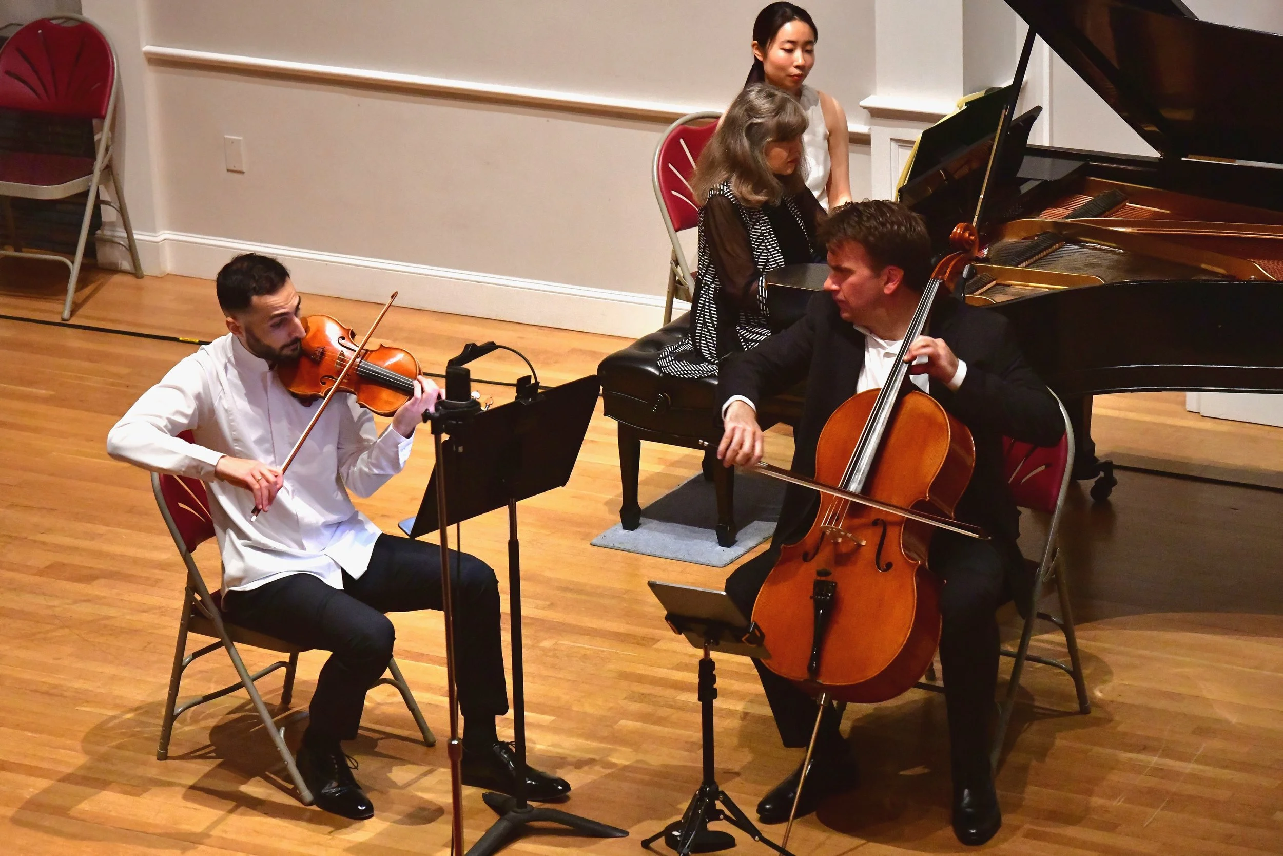 A string quartet performing on stage with a piano in the background. The ensemble includes a violinist, a cellist, and two women, one at the piano and one standing beside it.