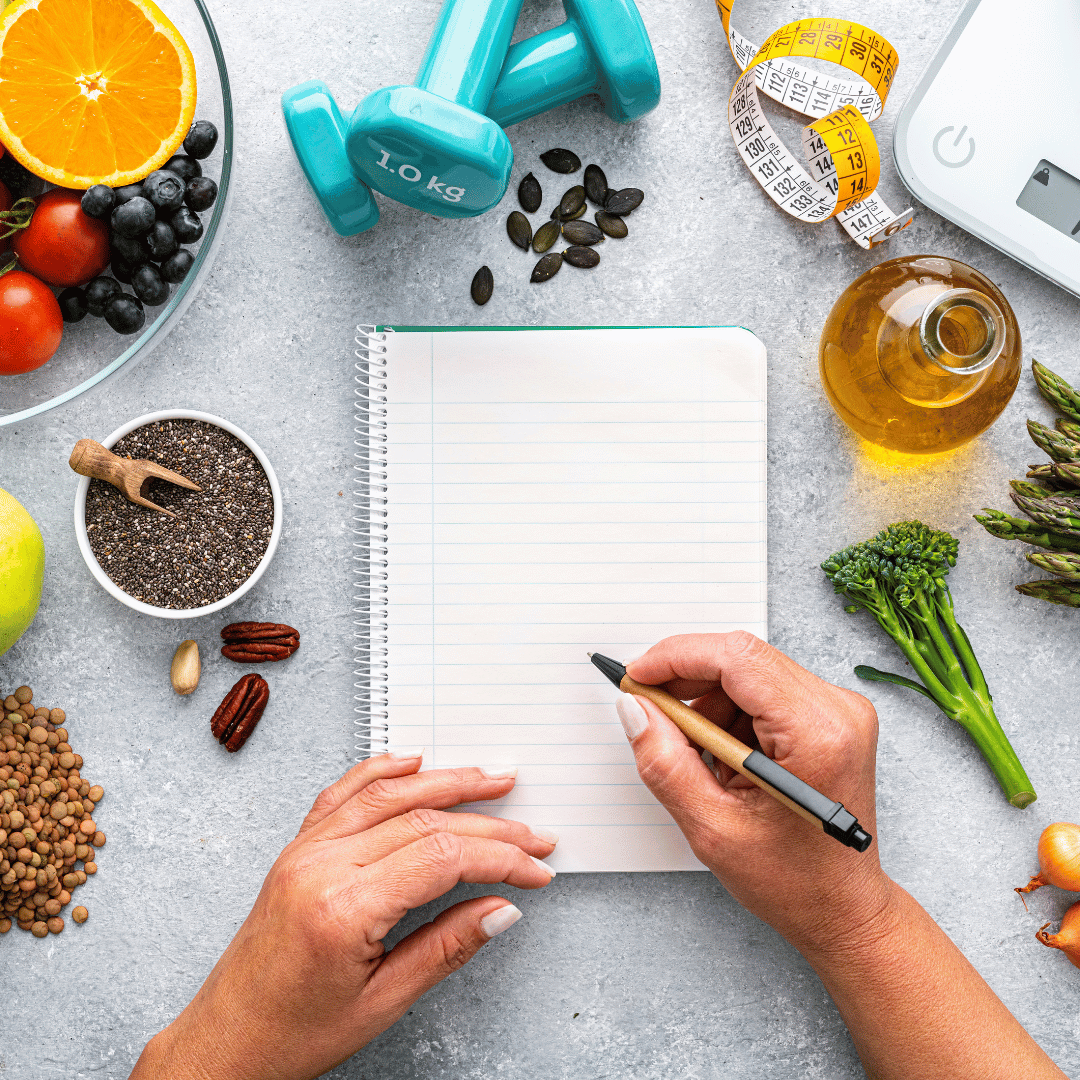 A person writing in a blank notebook surrounded by healthy foods, workout equipment, a scale, measuring tape, and other fitness-related items on a gray surface.