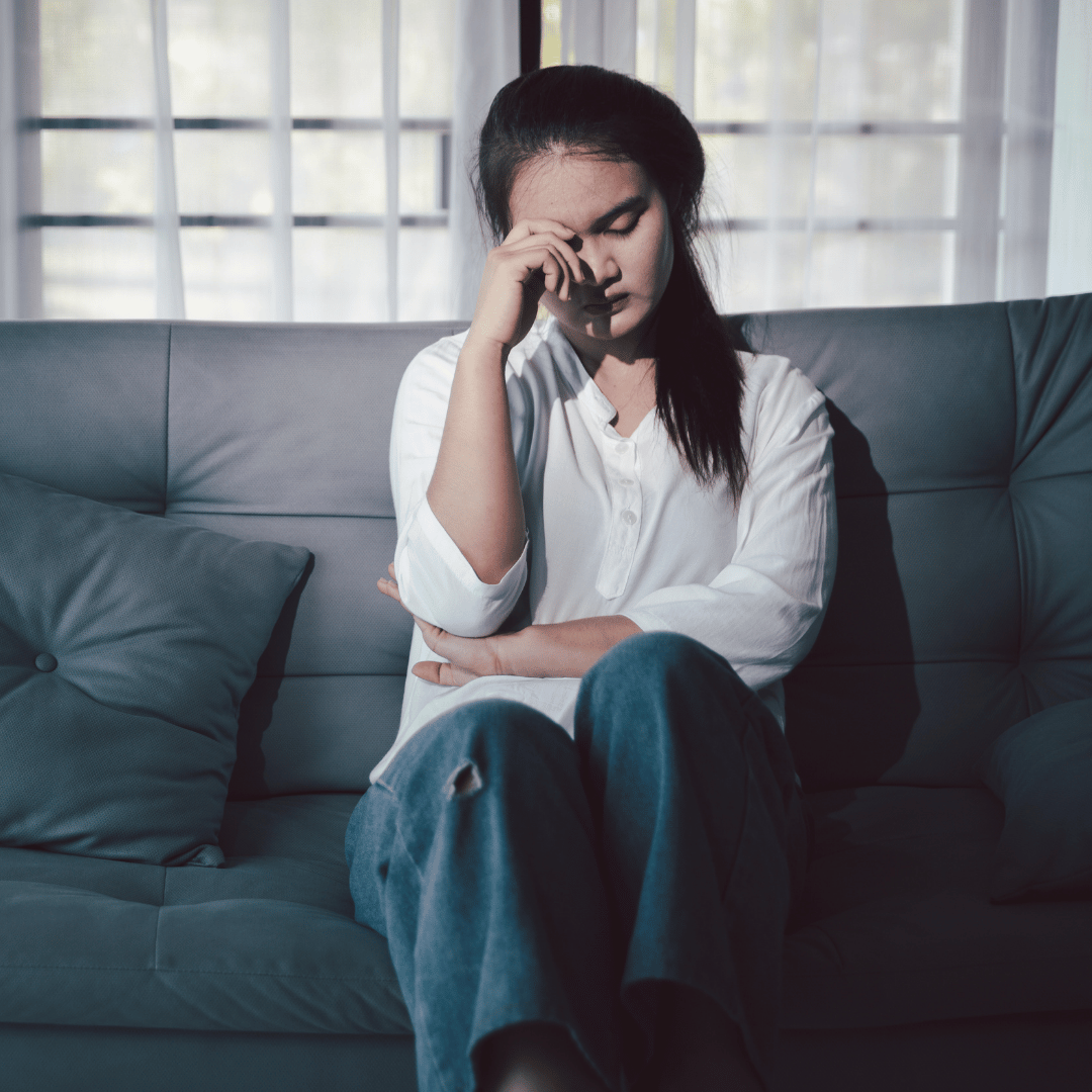 Young woman sitting on a gray couch with her eyes closed and hand on her forehead, appearing upset or tired, in front of a window with sheer curtains.
