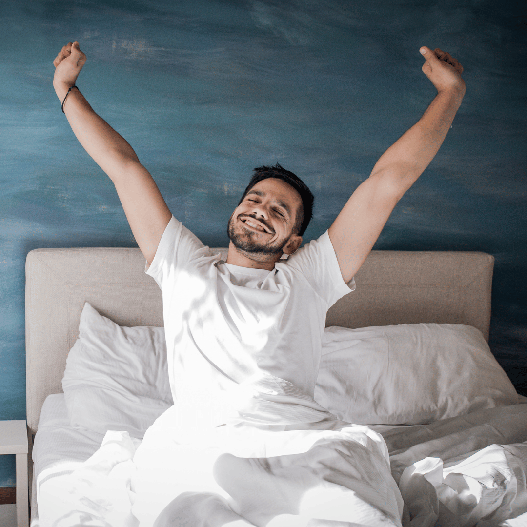 A man sitting up in bed, stretching and smiling with arms raised, in a bedroom with a blue wall and white bedding.