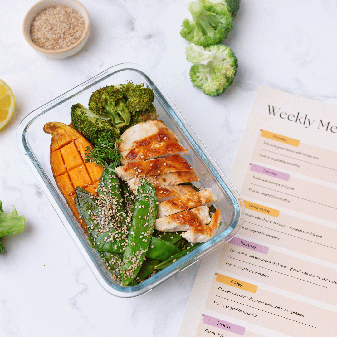 A glass container with sliced grilled chicken, broccoli, roasted sweet potato, snap peas topped with sesame seeds, and a garnish of herbs, placed on a white surface next to a weekly meal planner and a slice of lemon.