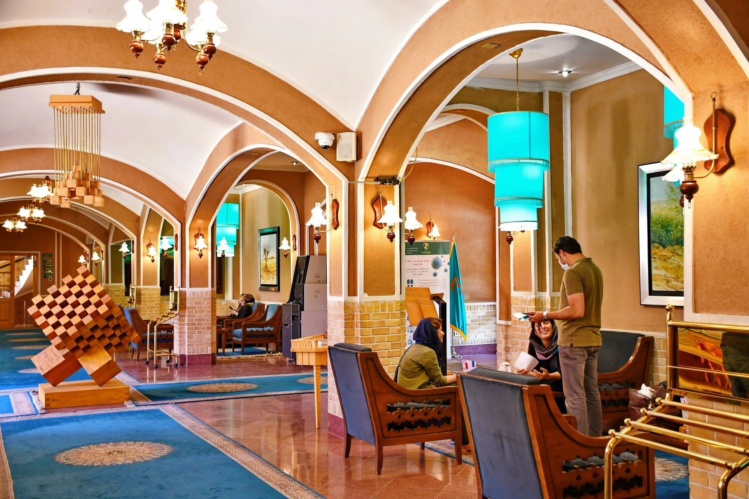 Interior of a lobby with arched ceilings, chandeliers, and seating areas. People are sitting and standing, with some wearing masks. There is a large wooden chessboard sculpture on the floor, and colorful hanging lights.