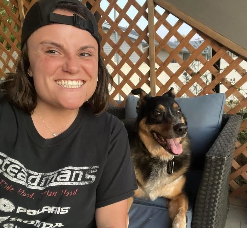 A young woman with brown hair wearing a black baseball cap backwards and a black graphic T-shirt, smiling and taking a selfie with her dog, a medium-sized black and tan dog with a happy expression, sitting on a patio with a wooden lattice fence in th